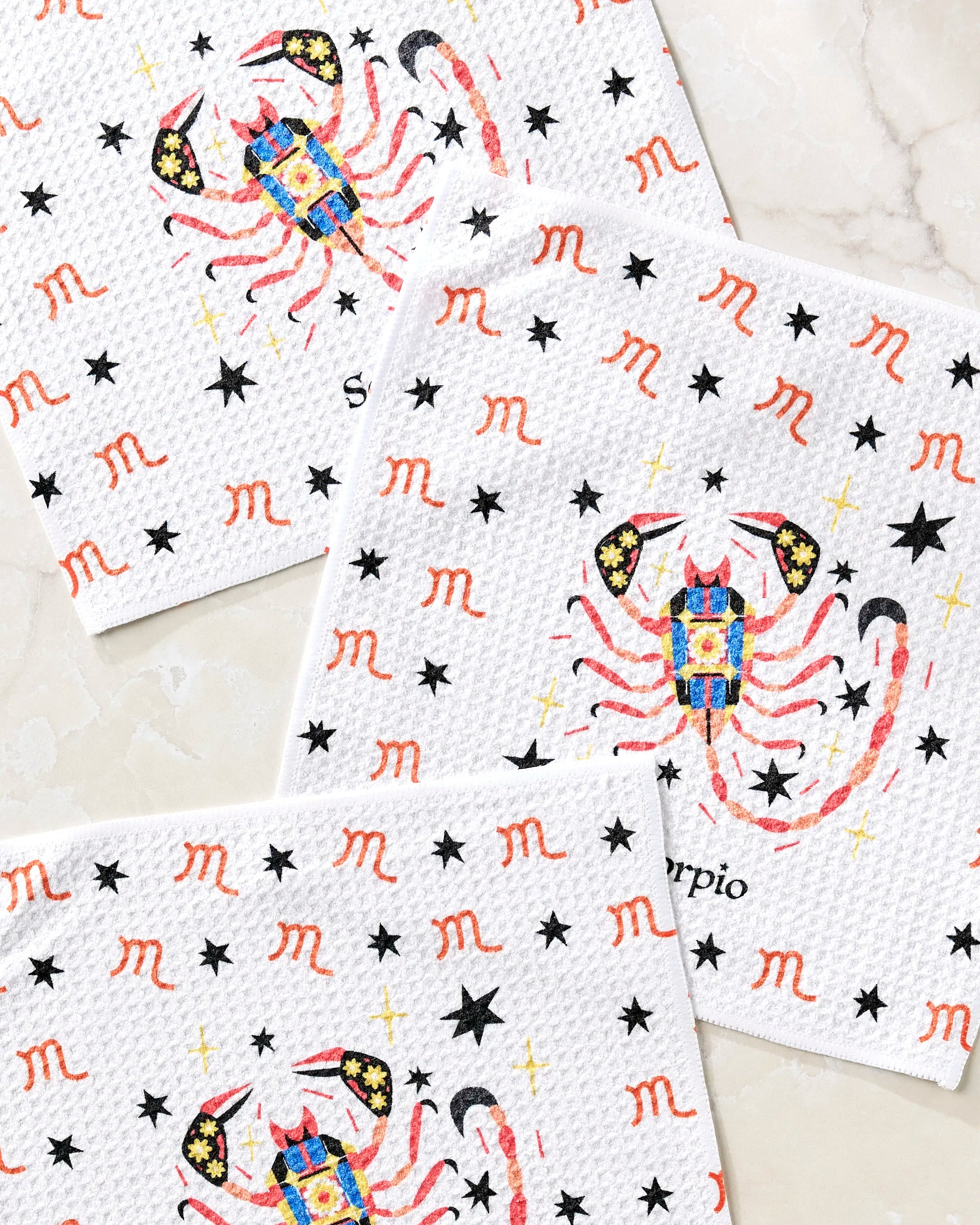 Three white "Mysterious Scorpio" dishtowels by Heather Zhou with colorful scorpions, black stars, red Scorpio zodiac symbols, and the word "Scorpio," displayed on a light marble surface.