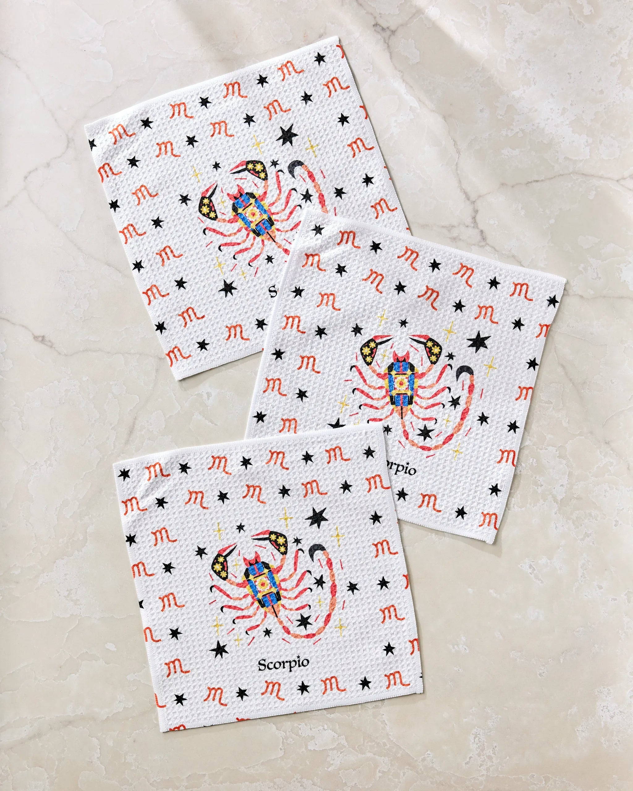Heather Zhou’s “Mysterious Scorpio” set includes three white napkins with red Scorpio zodiac signs, black stars, a vibrant scorpion in the center, and "Scorpio" text. Made from recycled materials and displayed on a light marble surface.