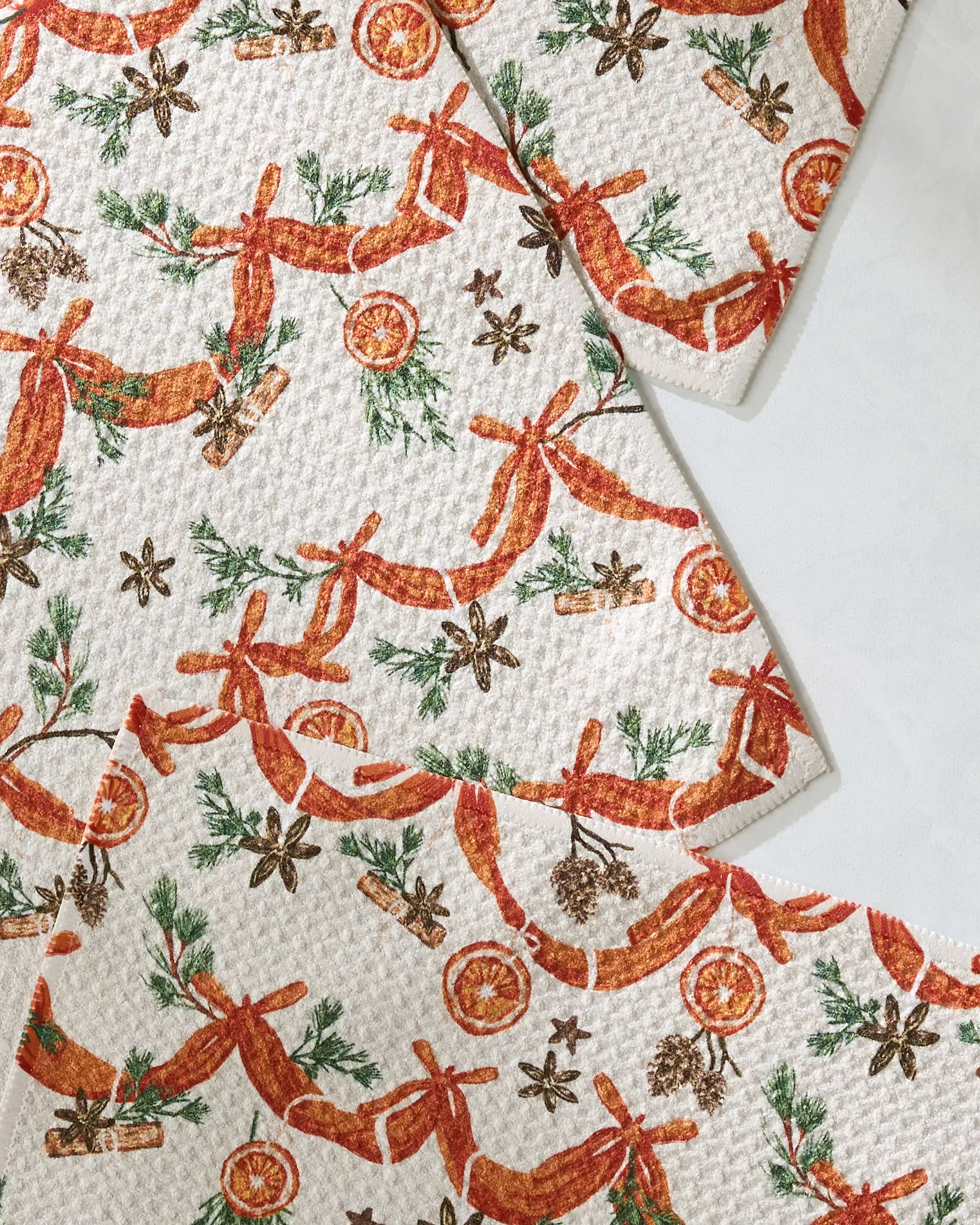 The Geometry Naranjas paper towels are made from recycled materials and feature festive brown and orange patterns of dried orange slices, cinnamon sticks, pinecones, and ribbons on a white background.