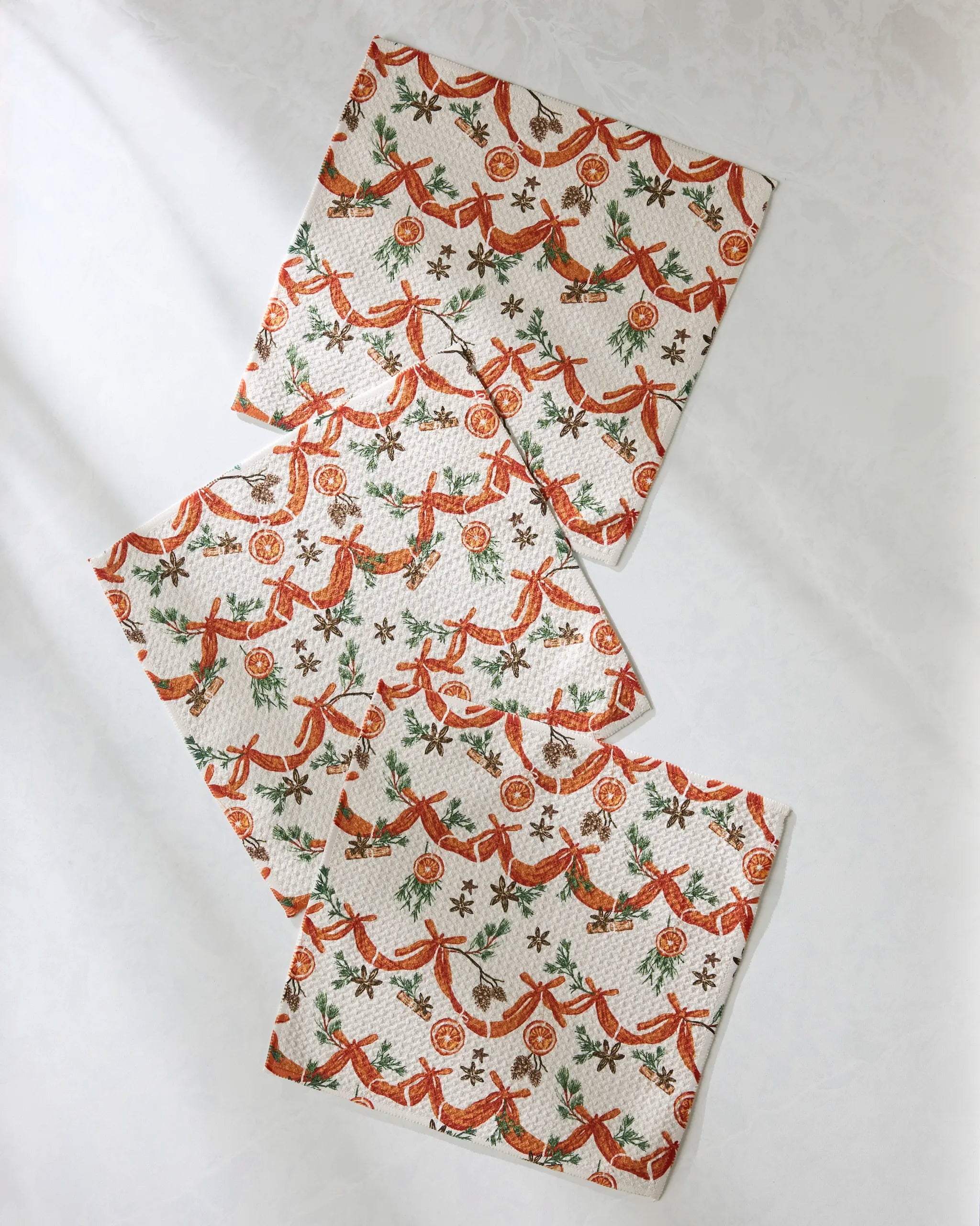 Three "Naranjas" napkins by Geometry, crafted from recycled materials, display a festive pattern of red birds, pine branches, and orange slices, arranged in a staggered layout on a white surface.