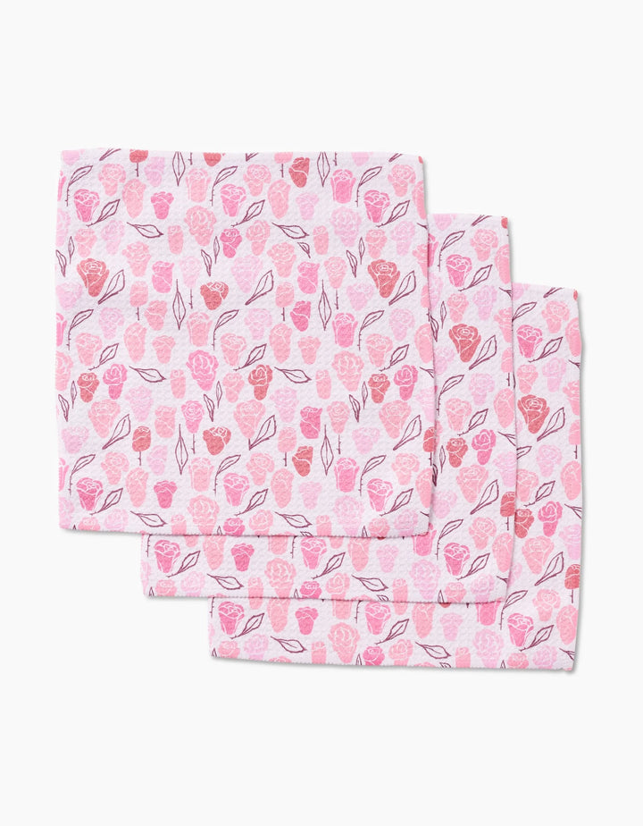Three folded Elisabeth Wing Neutral Rose cloth napkins, made from Geoweave fabric, display hand-drawn pink and red roses and leaves on a light pink background, stacked with slight overlap.