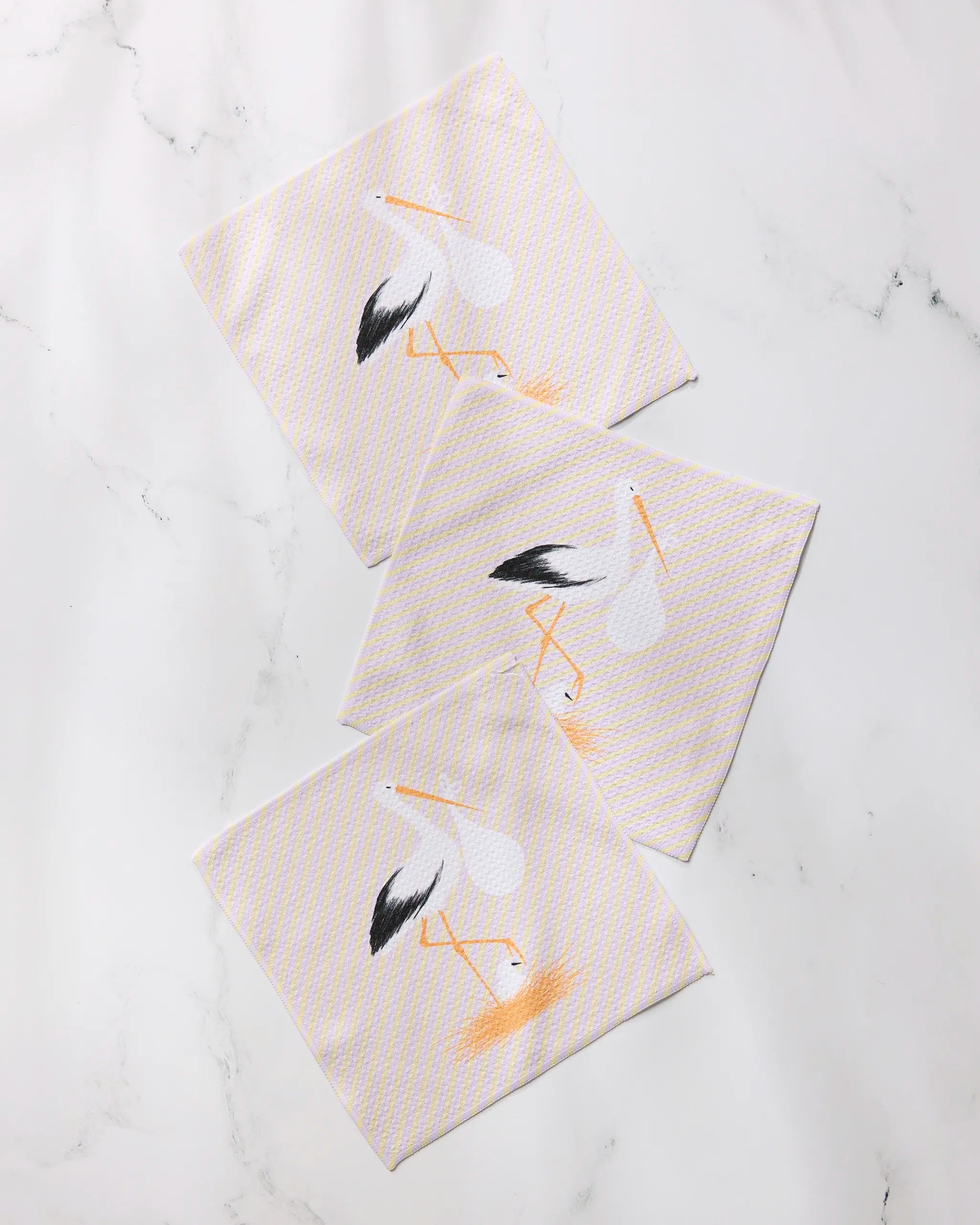 Geometry’s “New Mom” set includes three cream-colored recycled cloth napkins with a subtle checkered pattern and a stork illustration, displayed on white marble.