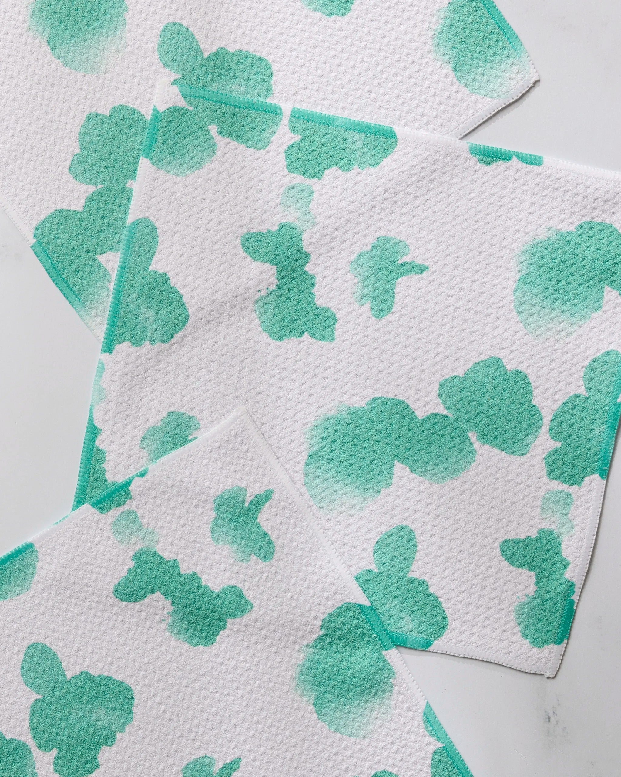 Four white Niamh kitchen towels by Geometry, featuring light green abstract cloud-like patterns, are overlapped on a white surface to showcase their absorbent textured design.