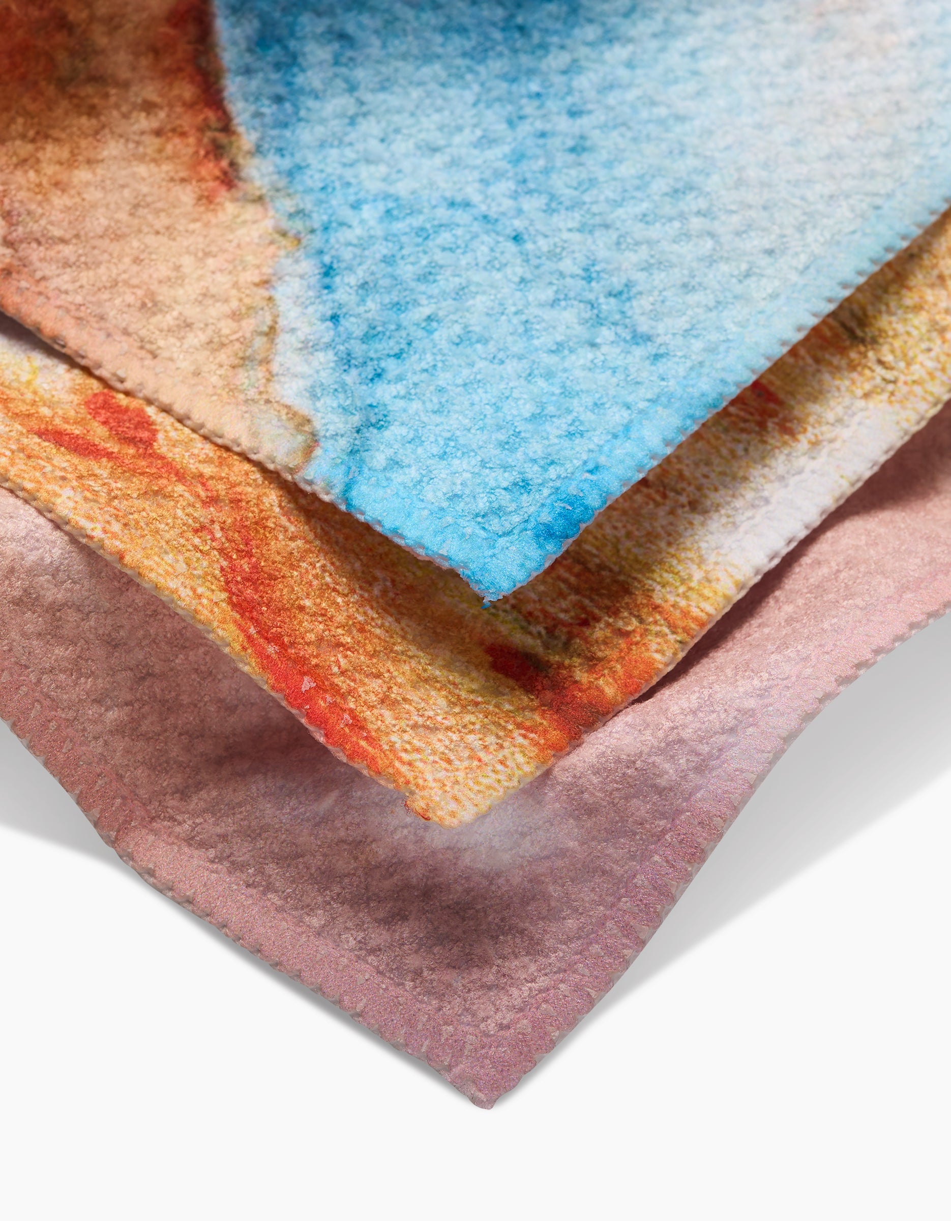 A close-up of three GEOMETRY Nice Water fabric pieces made from recycled materials shows pink, orange, and blue abstract watercolor patterns stacked neatly with textured surfaces and slightly visible edges.