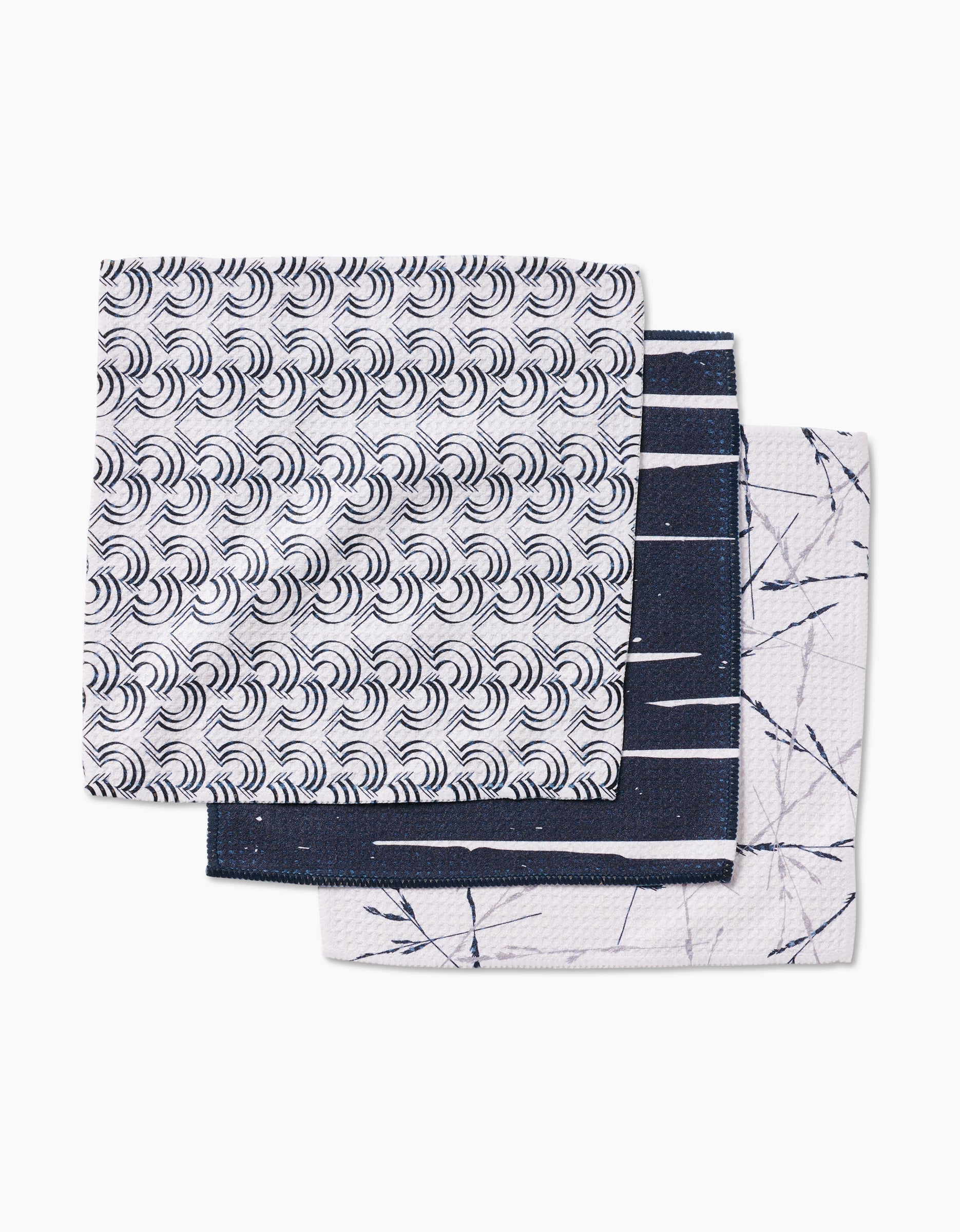 Three folded GEOMETRY dishcloths from the Night Reeds set are stacked: the top with a wave pattern, the middle dark with white lines, and the bottom white with a navy-blue branch design—all made from super-absorbent recycled materials.