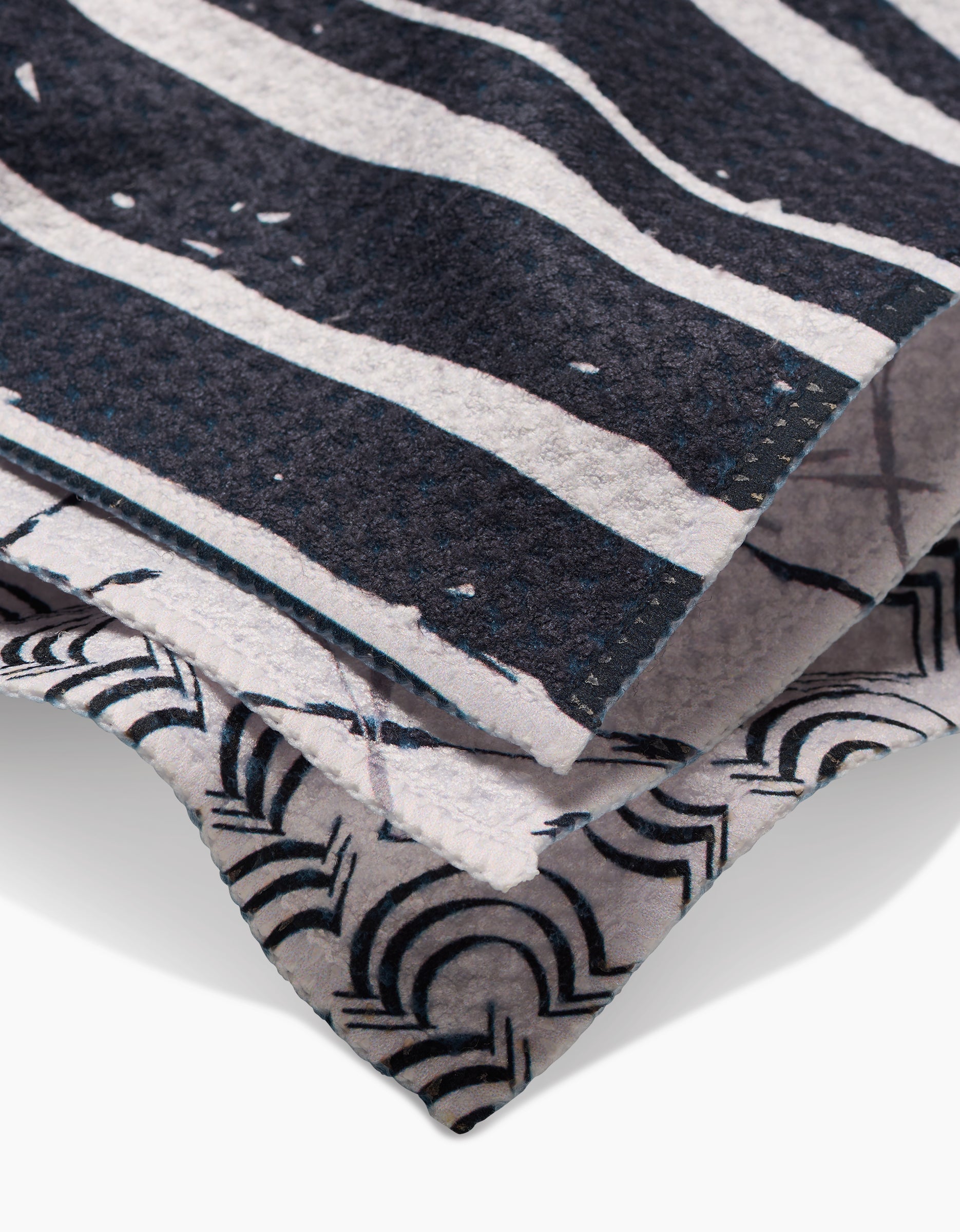 Close-up of GEOMETRYs Night Reeds dishcloths, featuring super-absorbent fabric with bold black and white wavy stripes and a geometric arc pattern, both showing textured surfaces and zigzag-cut edges.