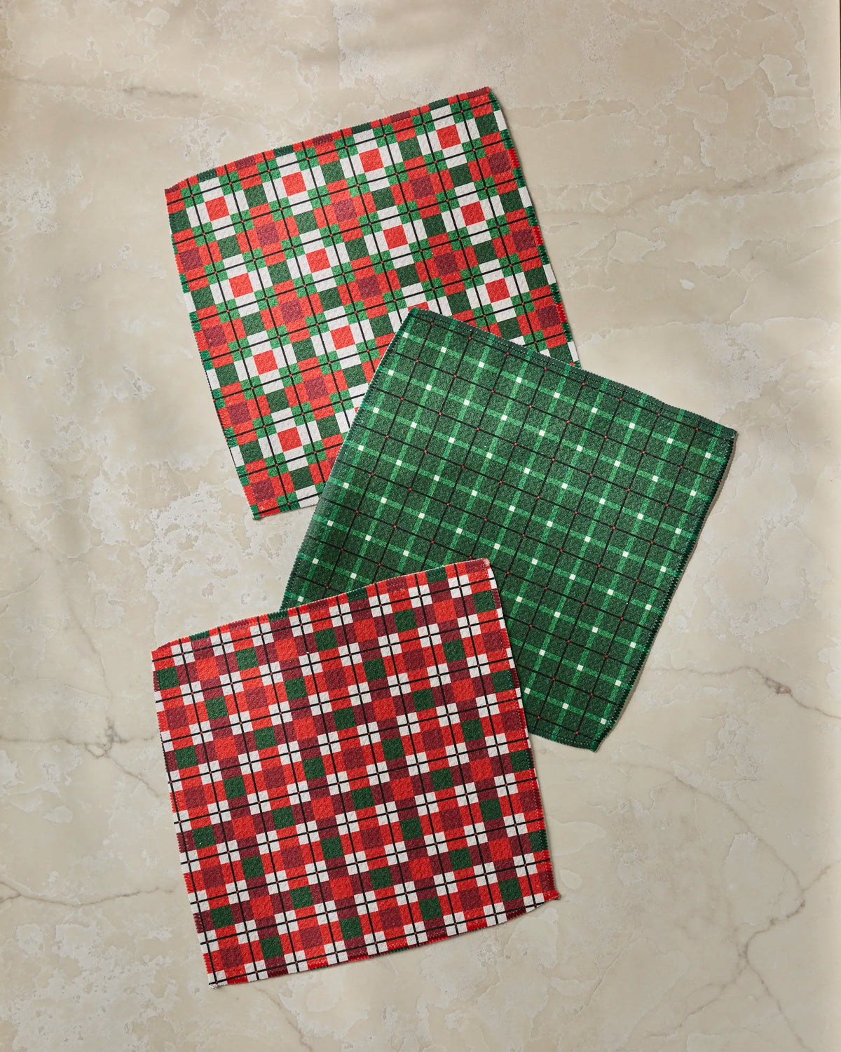Three folded cloth napkins from Geometry’s Noel Plaid set—featuring red and white, green and white, and red, green, and white plaid patterns—are arranged on a light marble surface, creating a charming dishcloth ensemble crafted from recycled materials.