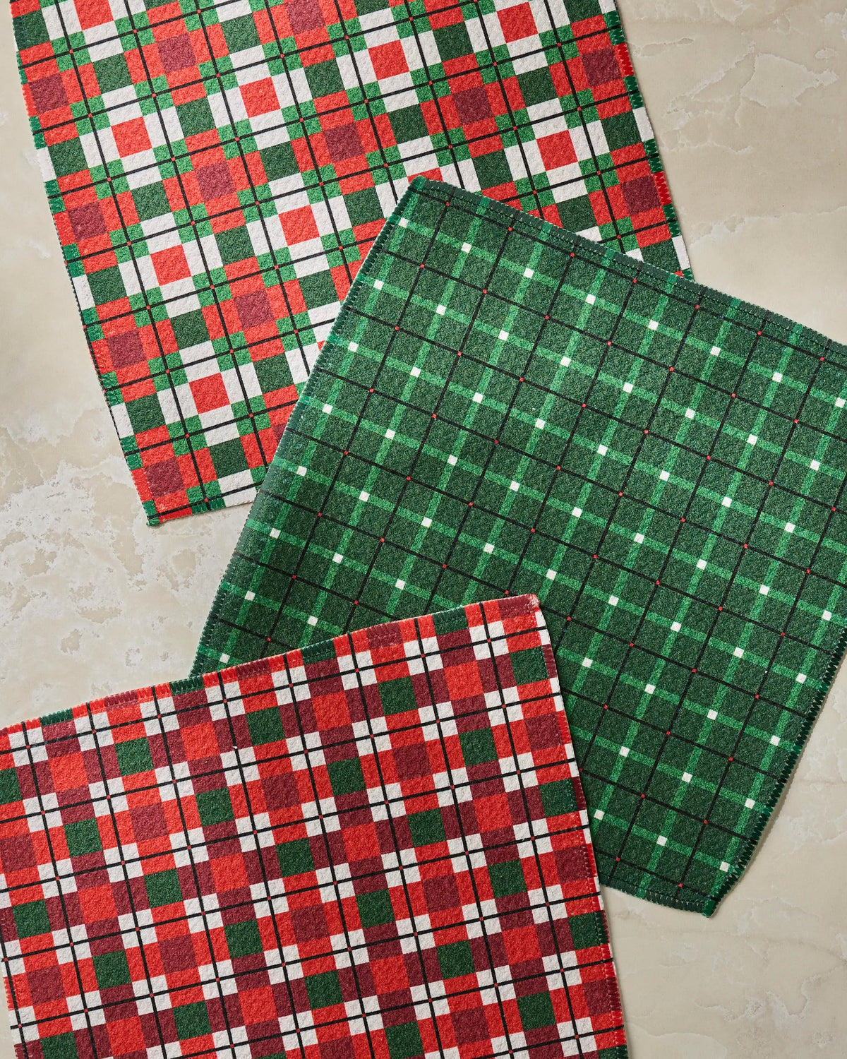 Three Noel Plaid cloth napkins from Geometry, featuring festive red, green, and white checker patterns, are arranged on a light beige marble surface. Made from recycled materials, this holiday-inspired set adds a classic touch to any kitchen décor.