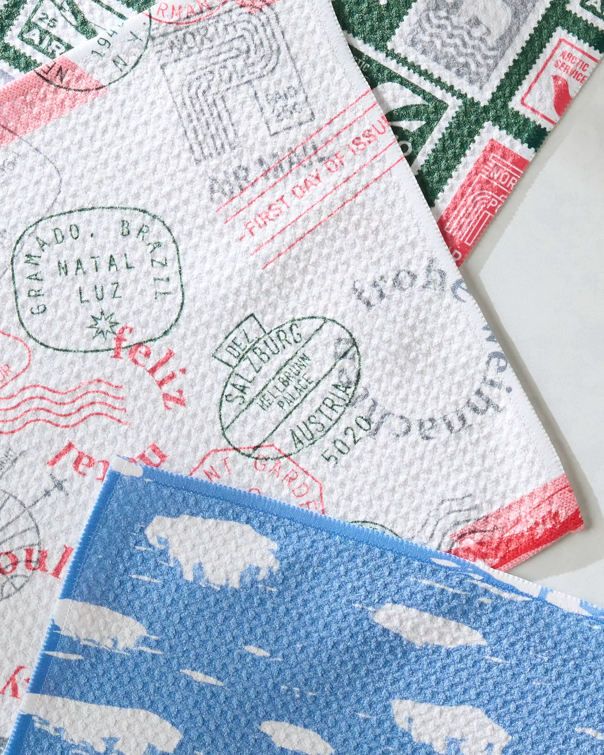 Three Nordic Cheer cloths by Geometry, made from recycled materials, overlap: one with green and red stamp prints, one with green stamps from Brazil and Austria, and a blue one featuring white buffalo silhouettes.