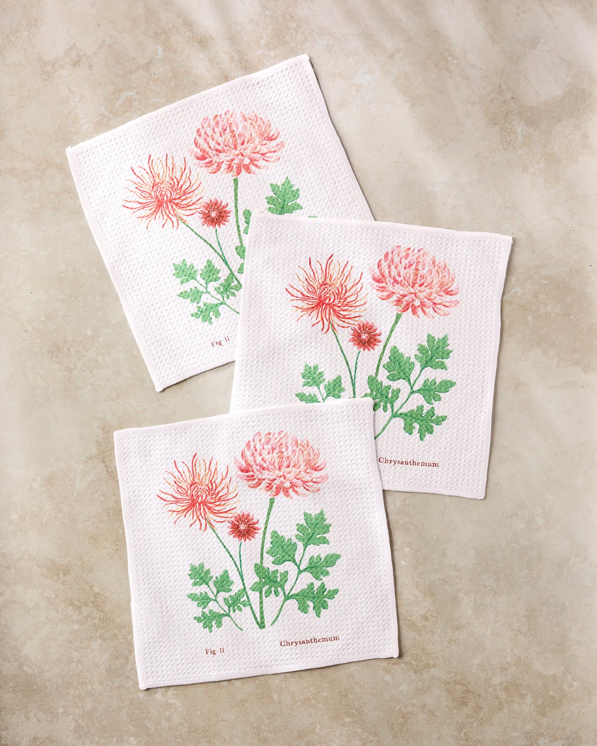 Three white "November Chrysanthemum" napkins by Linda Fox feature pink chrysanthemum flowers and green leaves, arranged on beige marble. Each is labeled “Chrysanthemum”—a charming addition to your absorbent dish towel set.