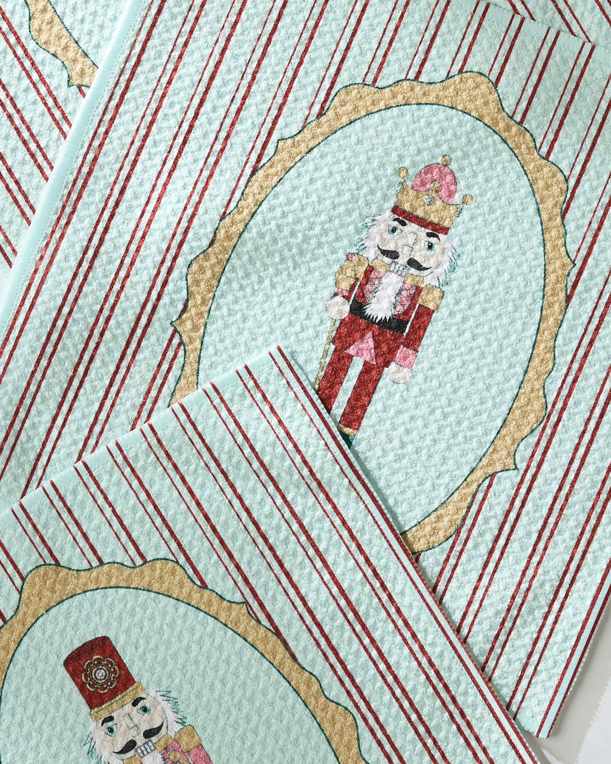 Geometry's Nutcracker Intermission dishtowels feature red stripes and nutcracker figures in gold ovals on a light blue textured background.