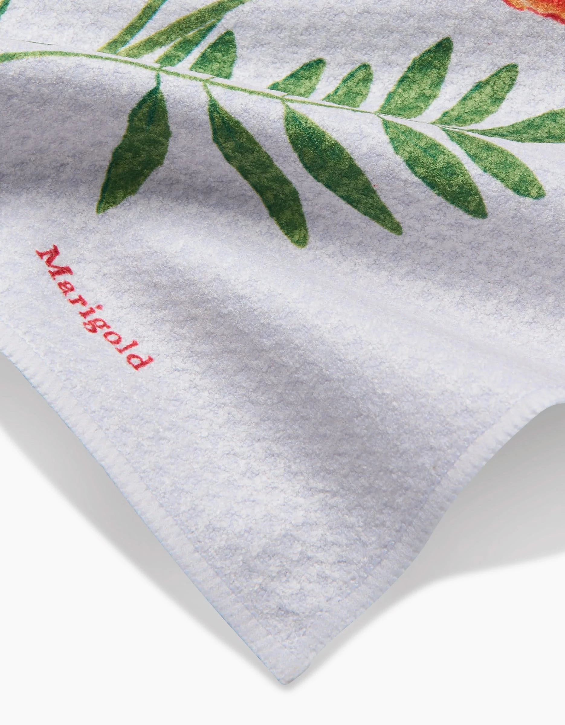 A close-up of a white Geoweave fabric towel by Linda Fox with a green leaf and October Marigold embroidered in red.