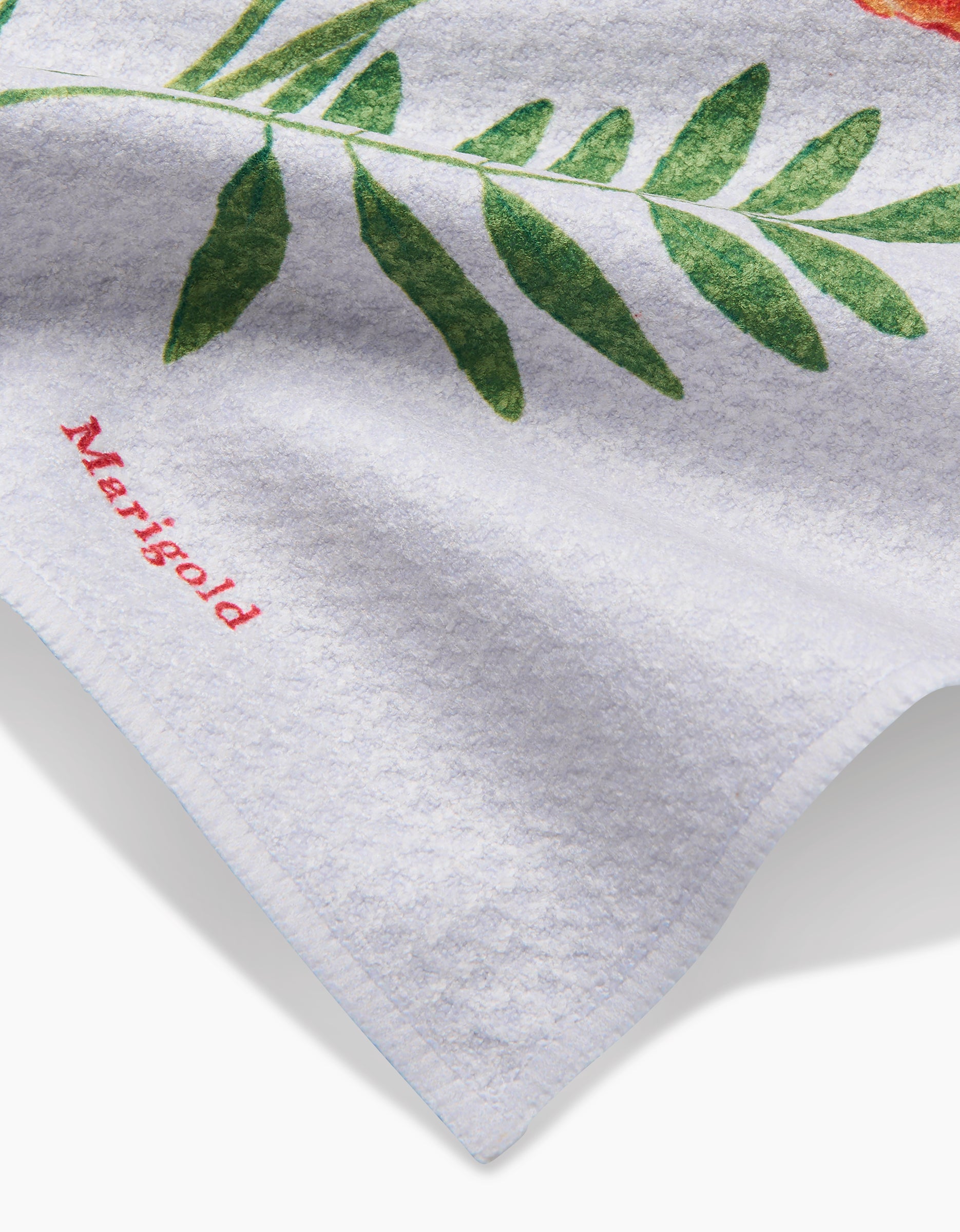 A close-up of a white Geoweave fabric towel by Linda Fox with a green leaf and October Marigold embroidered in red.