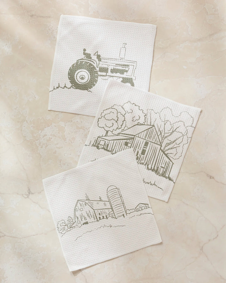Three Hannah Langford "Old Farm" white paper napkins—featuring simple line drawings of a tractor, barn with trees, and barn with silo—sit on cream marble, perfectly matching any kitchen dishcloth set or super-absorbent dishtowels.