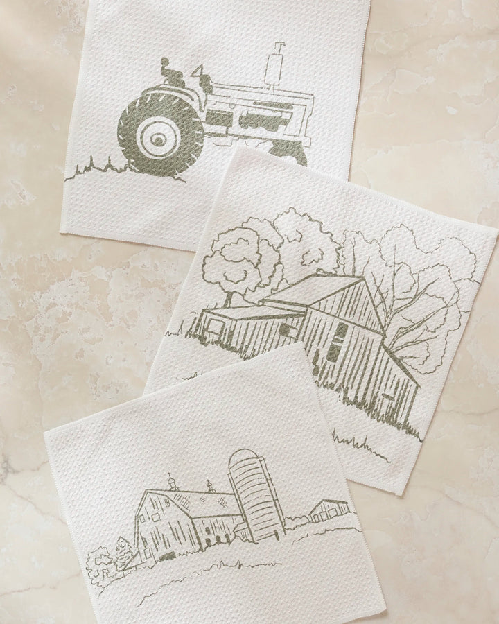 The Old Farm dishcloth set by Hannah Langford features three white napkins on a marble surface, each with a simple line drawing: a tractor, a barn with trees, and a barn with a silo—charming and practical for any kitchen.