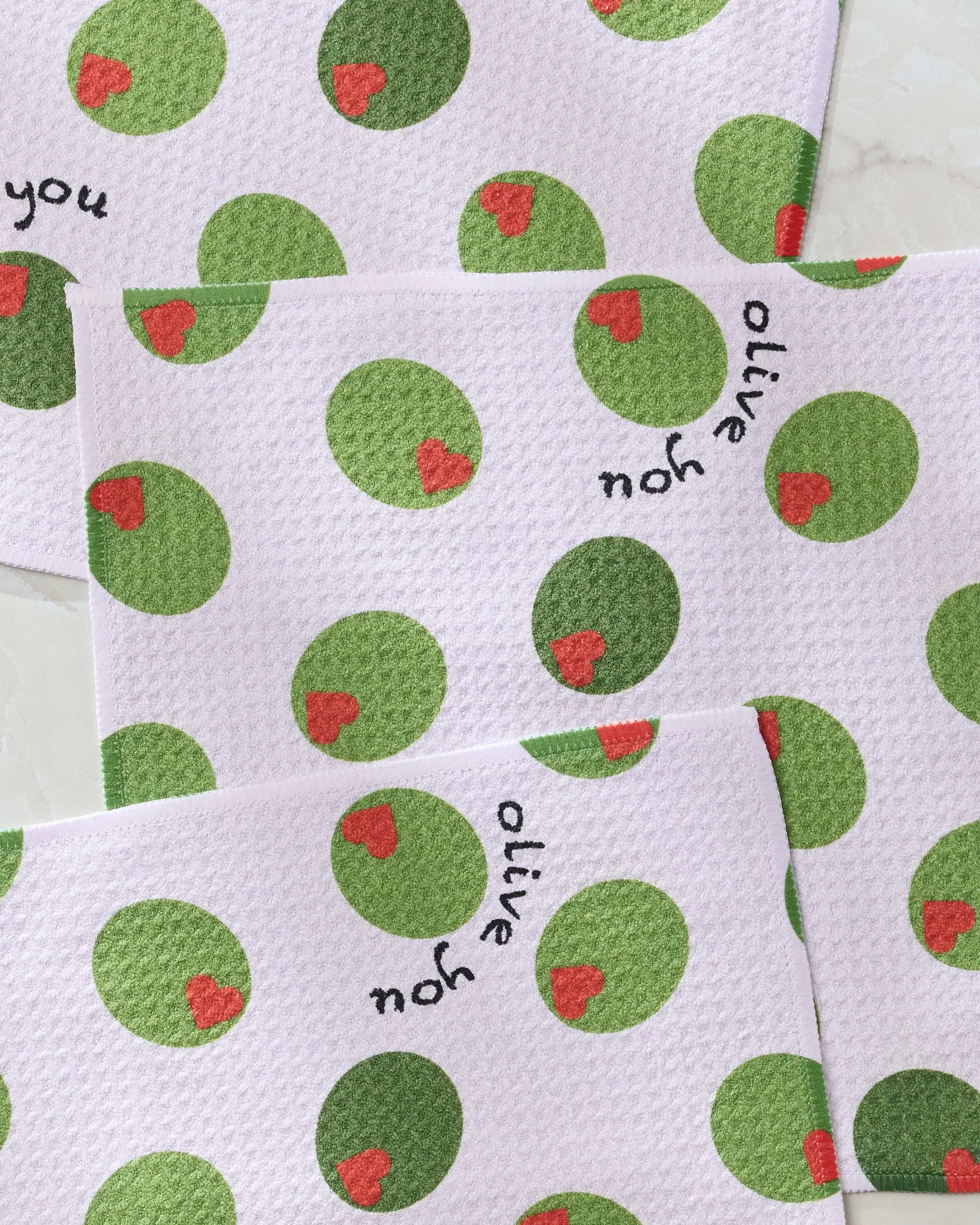 Three white, super-absorbent Geometry Olive You dishtowels feature green olive and red pimento patterns plus playful “olive you” text for a pun on “I love you.” This eco-friendly dishcloth set is shown on a light surface.