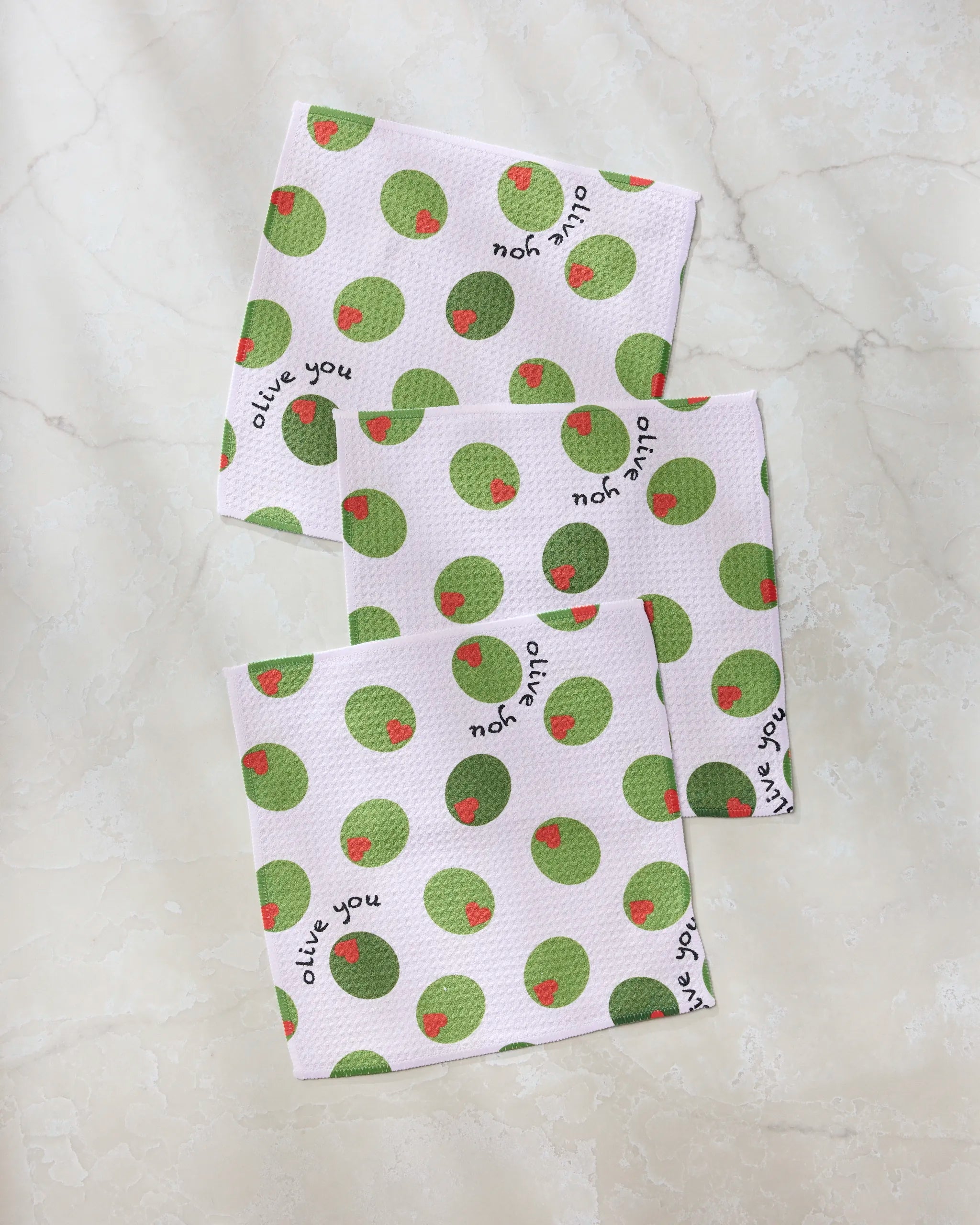 The Geometry Olive You dishcloth set includes three super-absorbent white cloths with a green olive pattern and "olive you" printed on each, beautifully arranged on a light marble surface—perfect for everyday use.