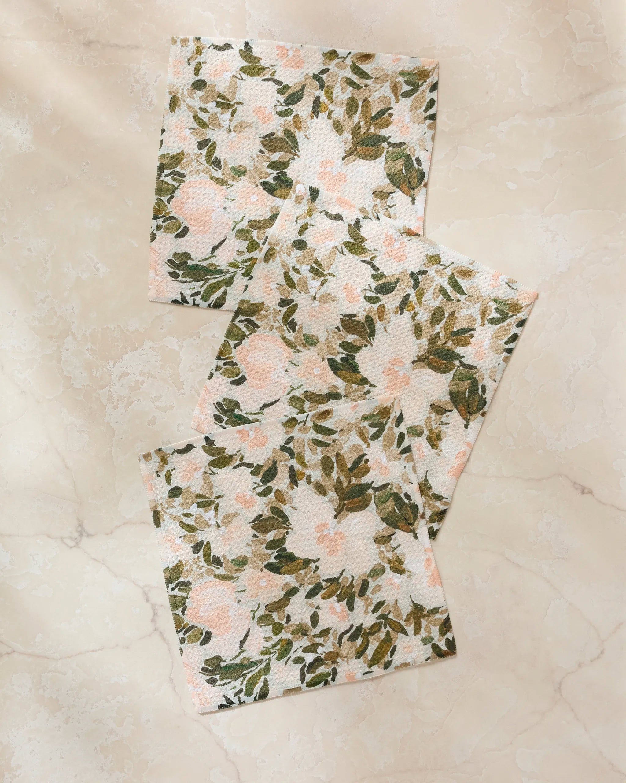 Three "Painted Blossoms" paper napkins by Anee Shah, with green leaf and pink floral patterns, are arranged on a light marble surface—an elegant touch to pair with your favorite absorbent dishcloths.