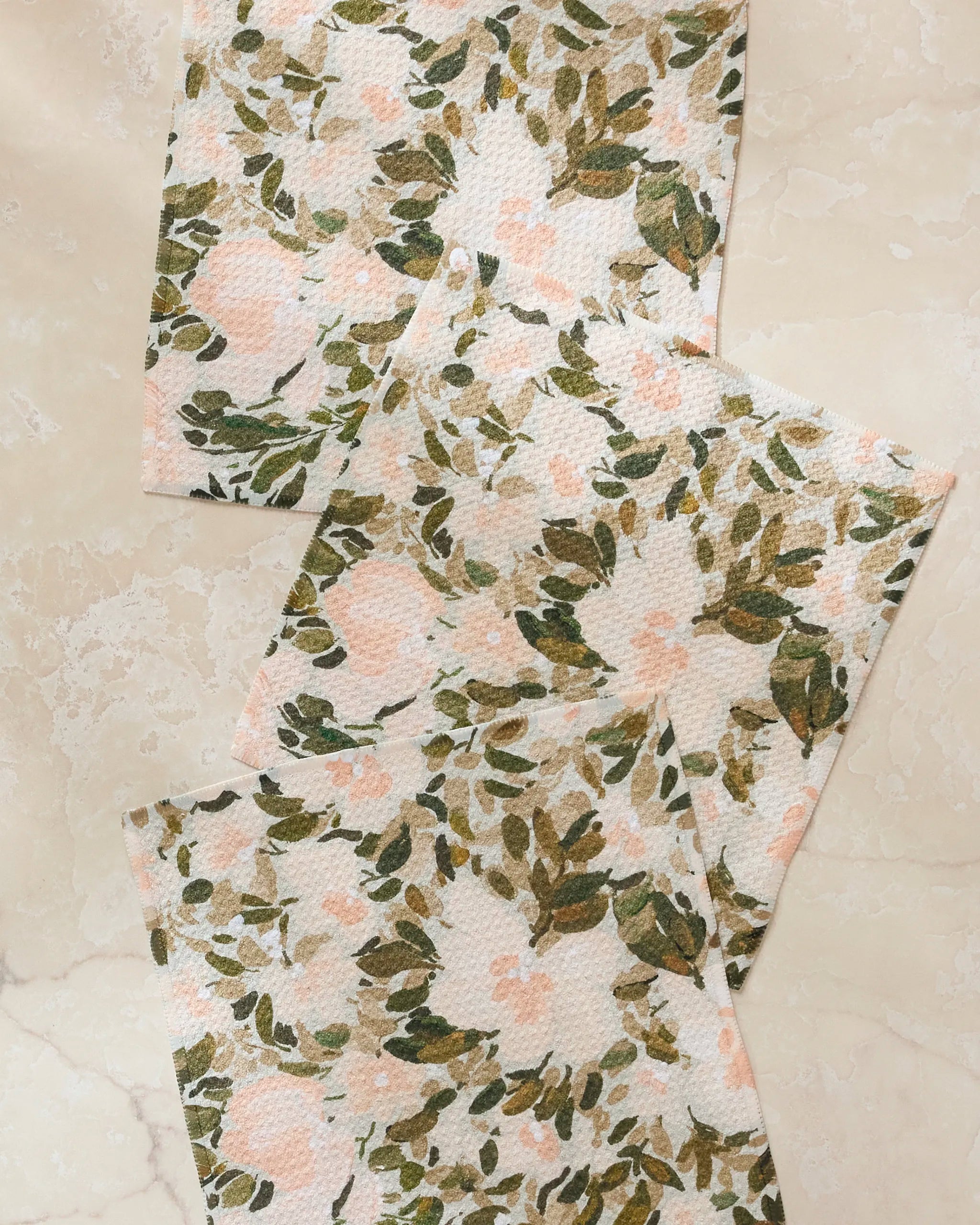 Three Painted Blossoms rectangular dishcloths by Anee Shah, featuring a green, olive, and pale pink floral pattern, are arranged slightly overlapping on a cream marble surface.