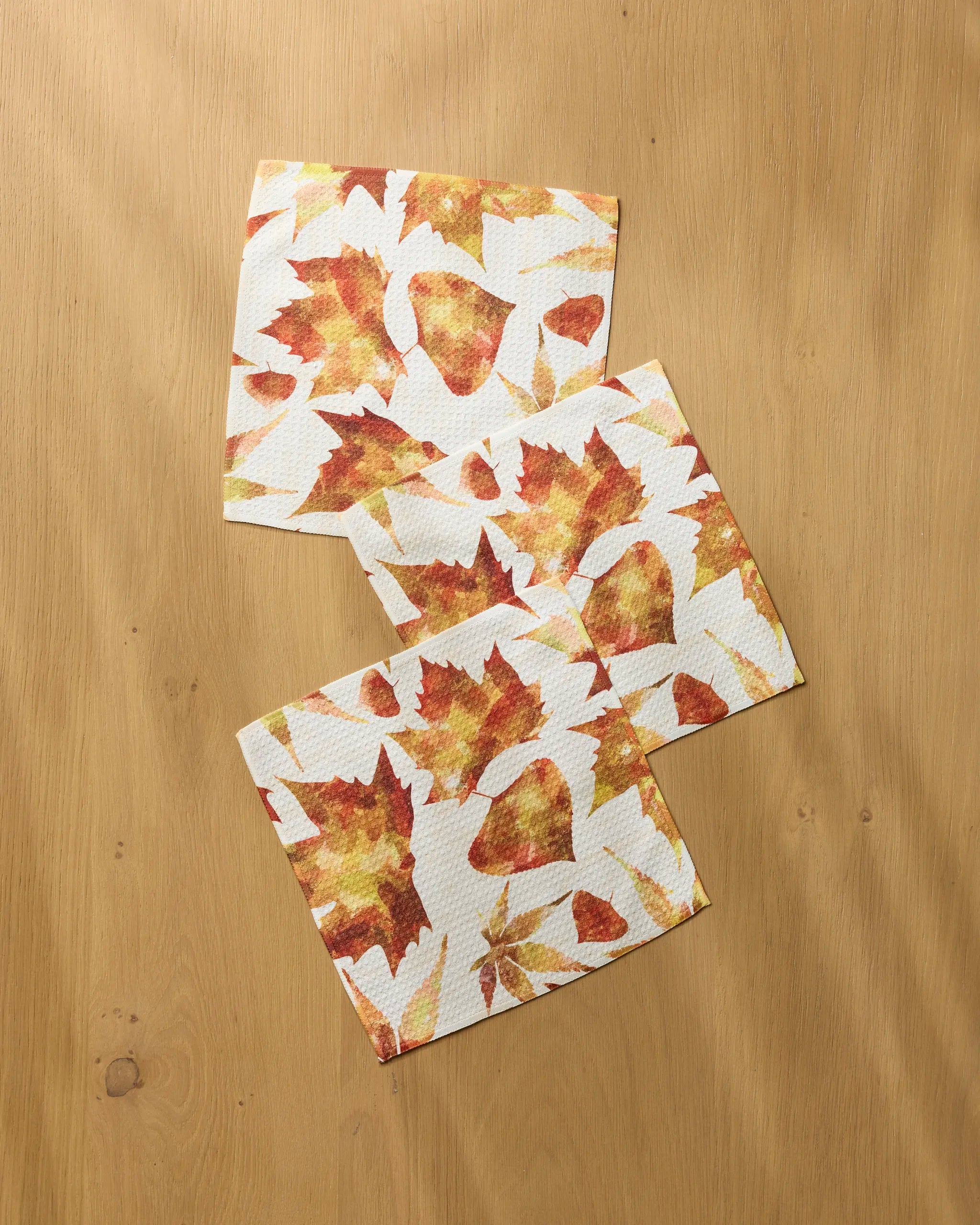 Three Geometry “Painted Fall” napkins featuring an autumn leaf pattern in orange, yellow, and brown are arranged on a light wooden surface for a seasonal display.