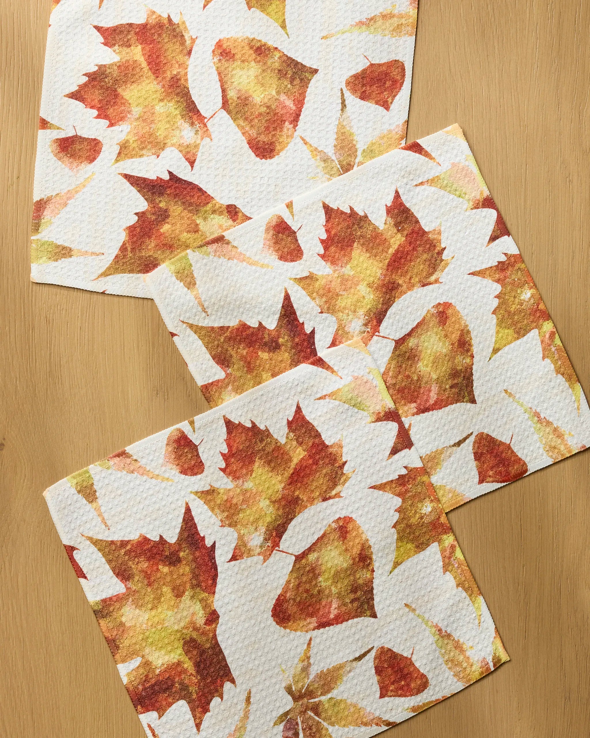 Three "Painted Fall" paper napkins by Geometry, made from recycled materials, showcase an autumn leaf pattern in orange, yellow, and brown with various shapes for a festive fall design on a wooden surface.