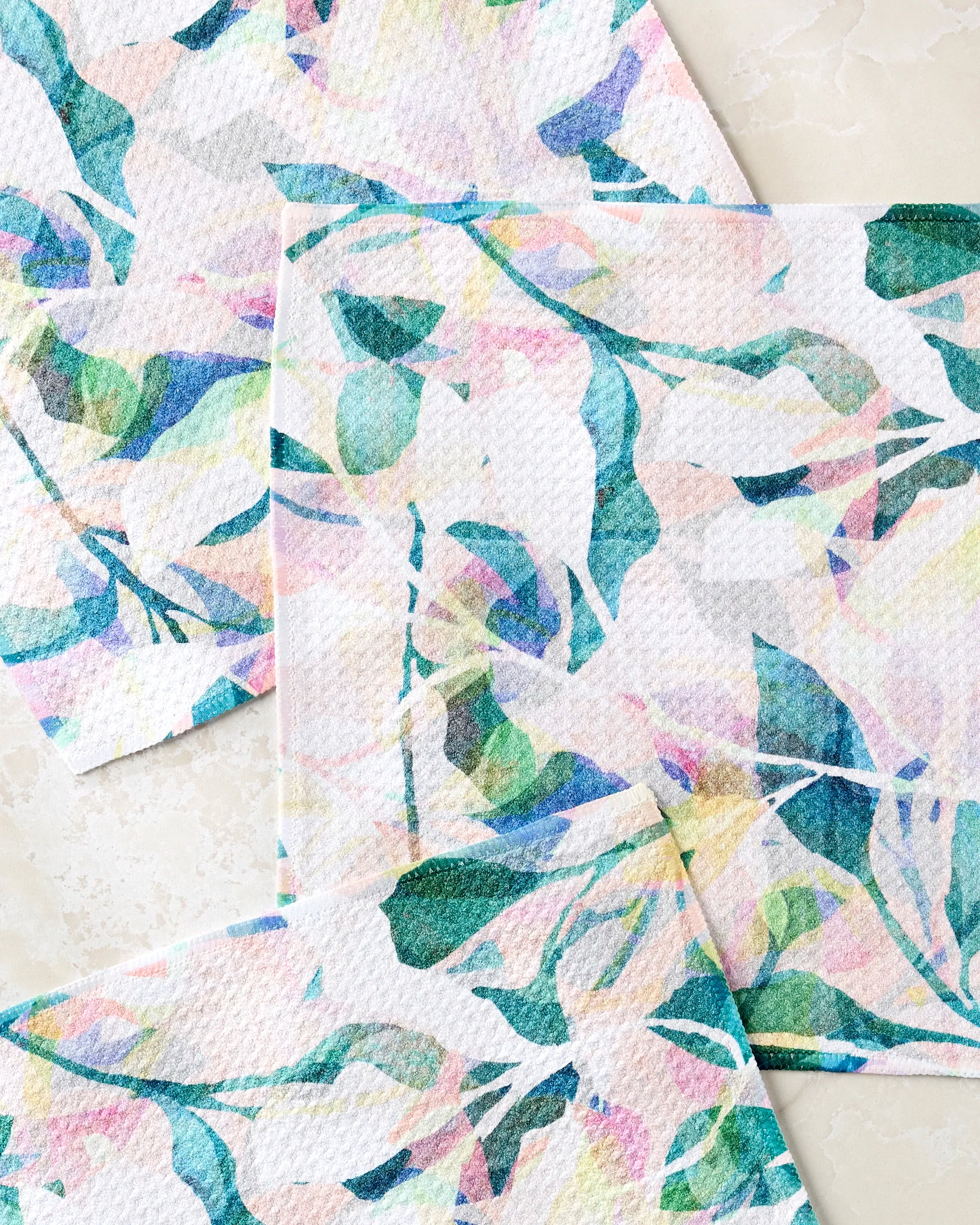Three "Painted Foliage" kitchen towels by Rachel Parker, featuring abstract leaf and branch patterns in blue, green, pink, and yellow on white recycled fabric, are arranged overlapping on a light marble surface.