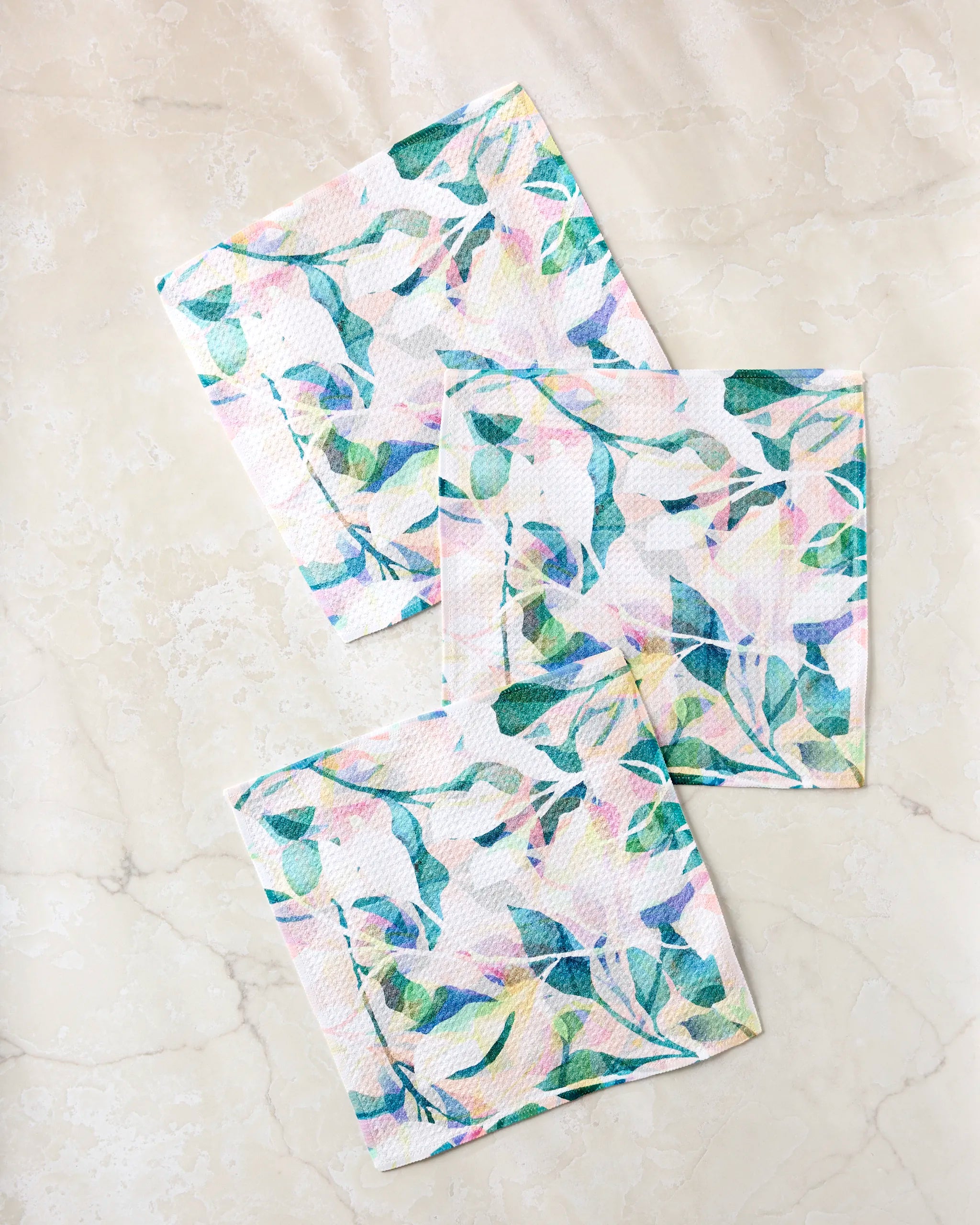 Three “Painted Foliage” square cloth napkins by Rachel Parker, featuring a pastel floral and leafy pattern, are casually overlapped on a light beige marble surface as part of an absorbent dishcloth set.