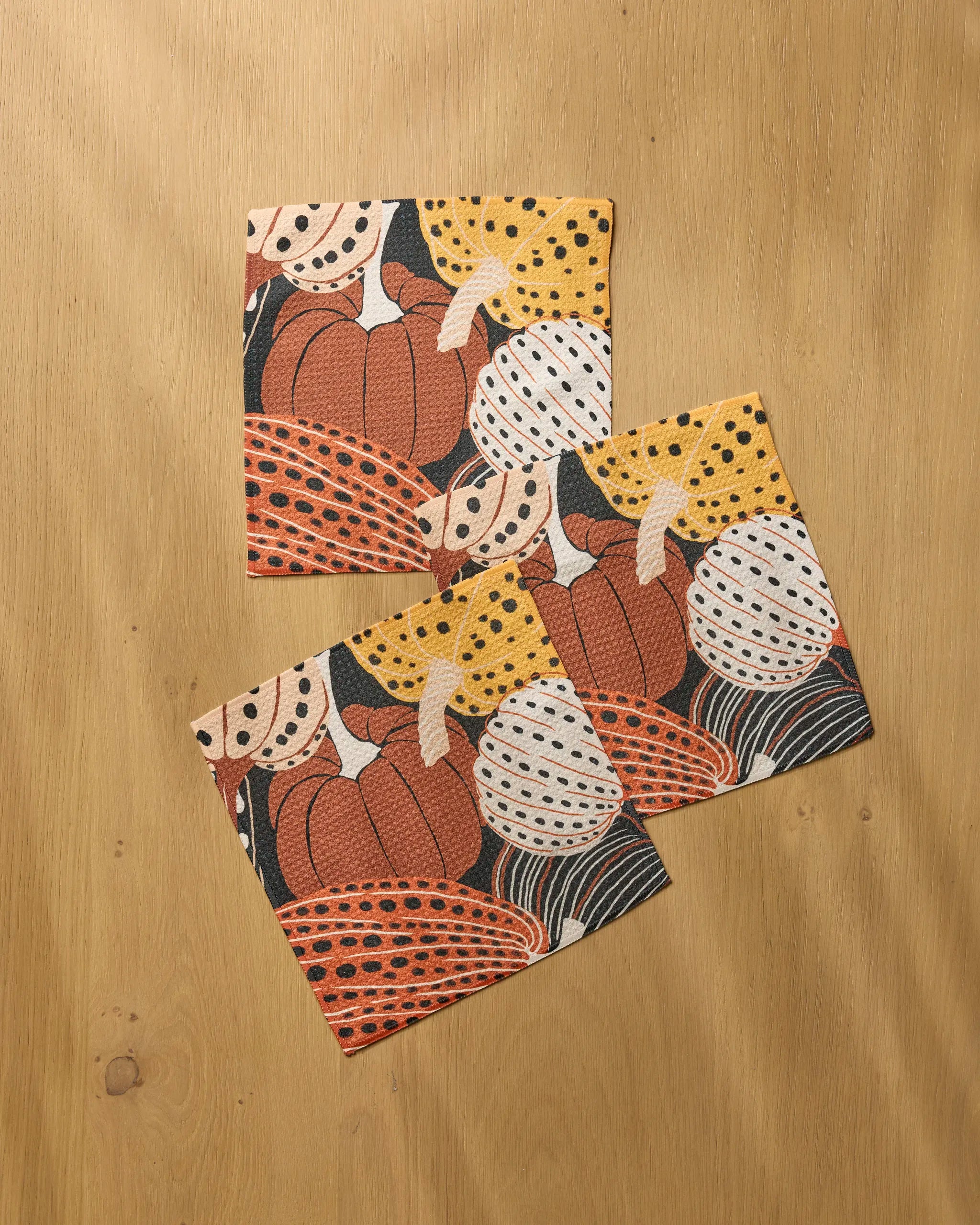 Three Painted Patch napkins by Geometry, made from recycled materials and featuring a vibrant pumpkin and gourd design in orange, yellow, white, and black, are displayed on a light wooden surface.