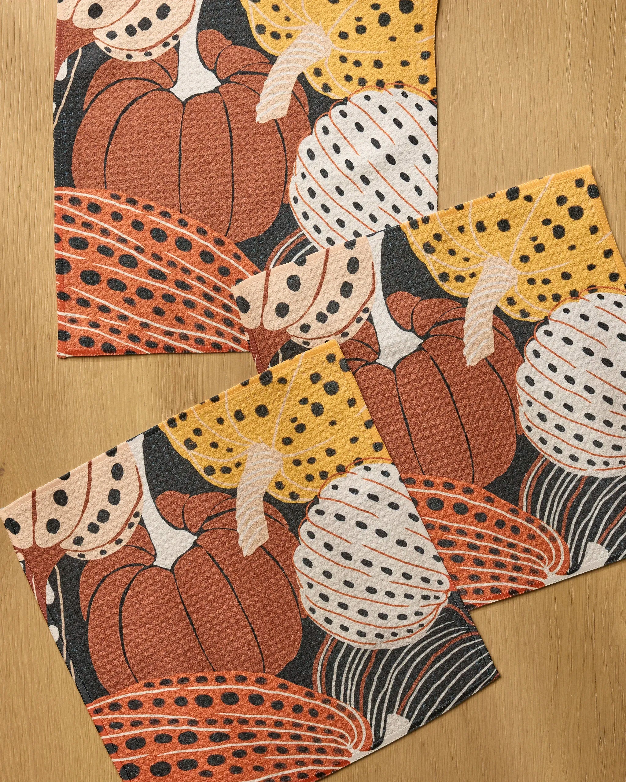 Three Painted Patch dishcloths by Geometry, featuring autumn-themed prints with pumpkins and gourds in orange, yellow, white, and black patterns on a wooden surface. Made from recycled materials for an eco-friendly touch.