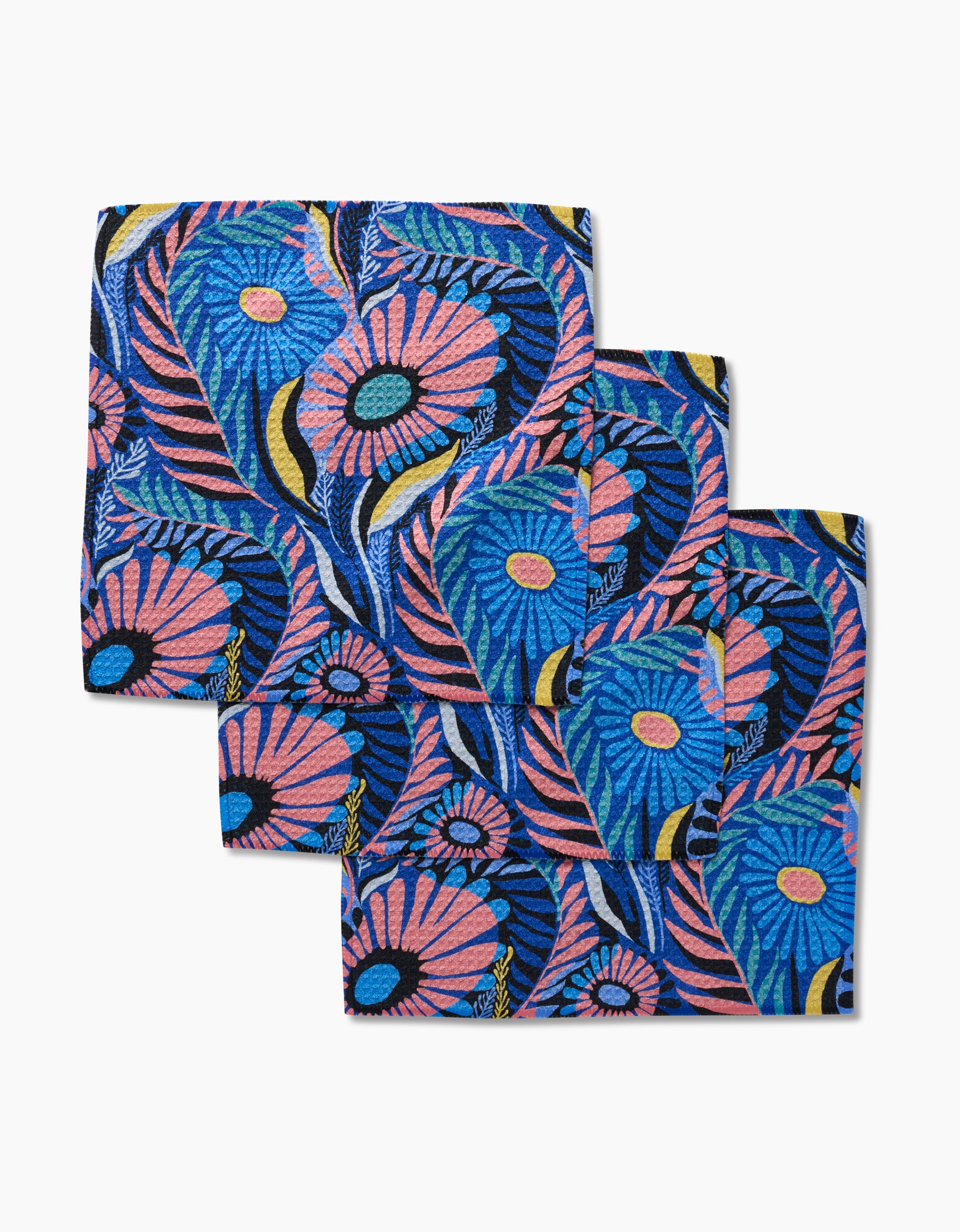Three folded Palm Beach fabric napkins by Masha Volnova, made from eco-friendly Geoweave, feature a vibrant floral pattern with large pink and orange flowers, blue leaves, and yellow accents on a dark background, overlapping on white.