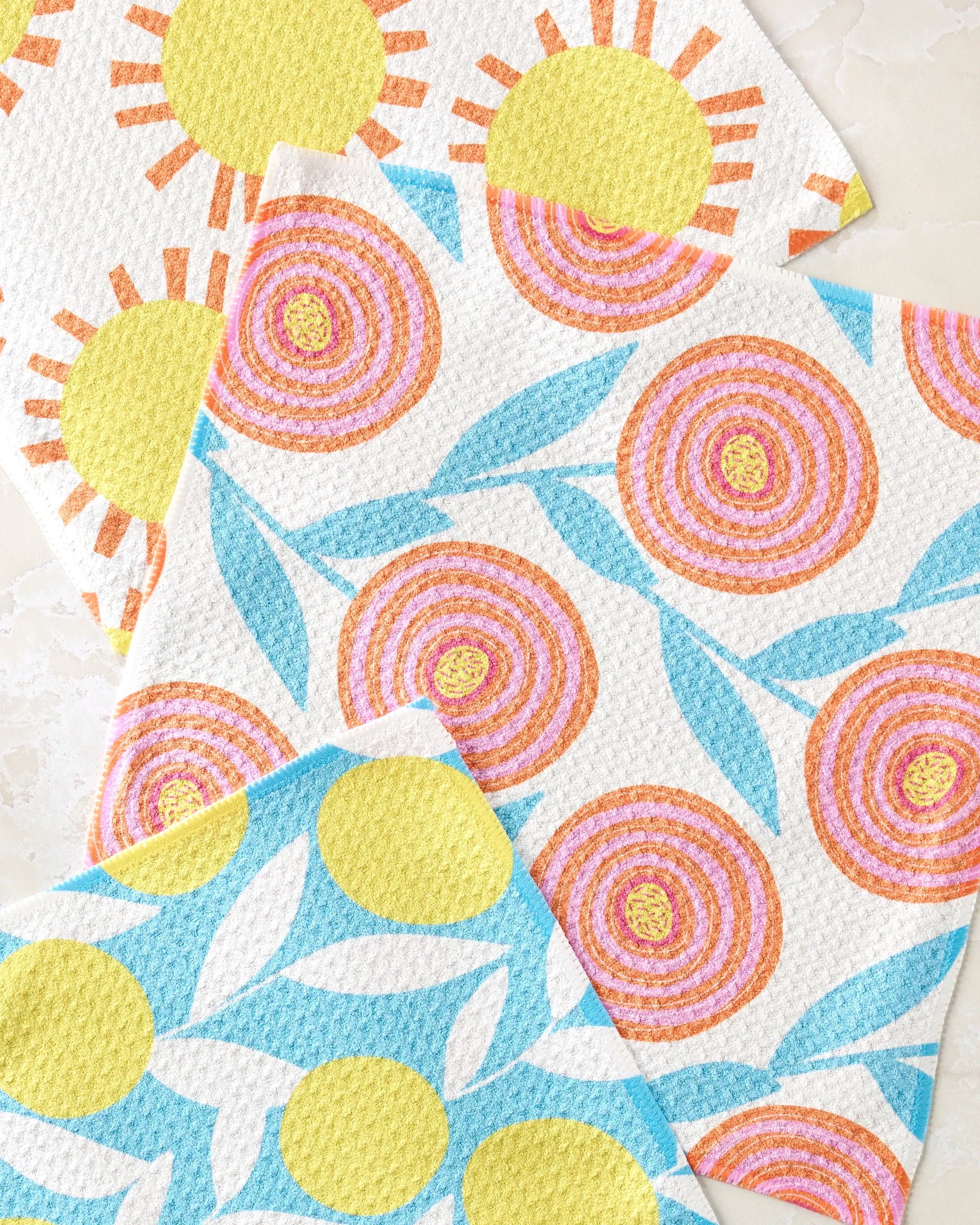 Parade by Lizzie Clark: a vibrant dishcloth set featuring yellow suns with orange rays, orange spirals with blue leaves, and yellow circles with white petals on blue—three bold recycled-fiber cloths overlap on a light background.