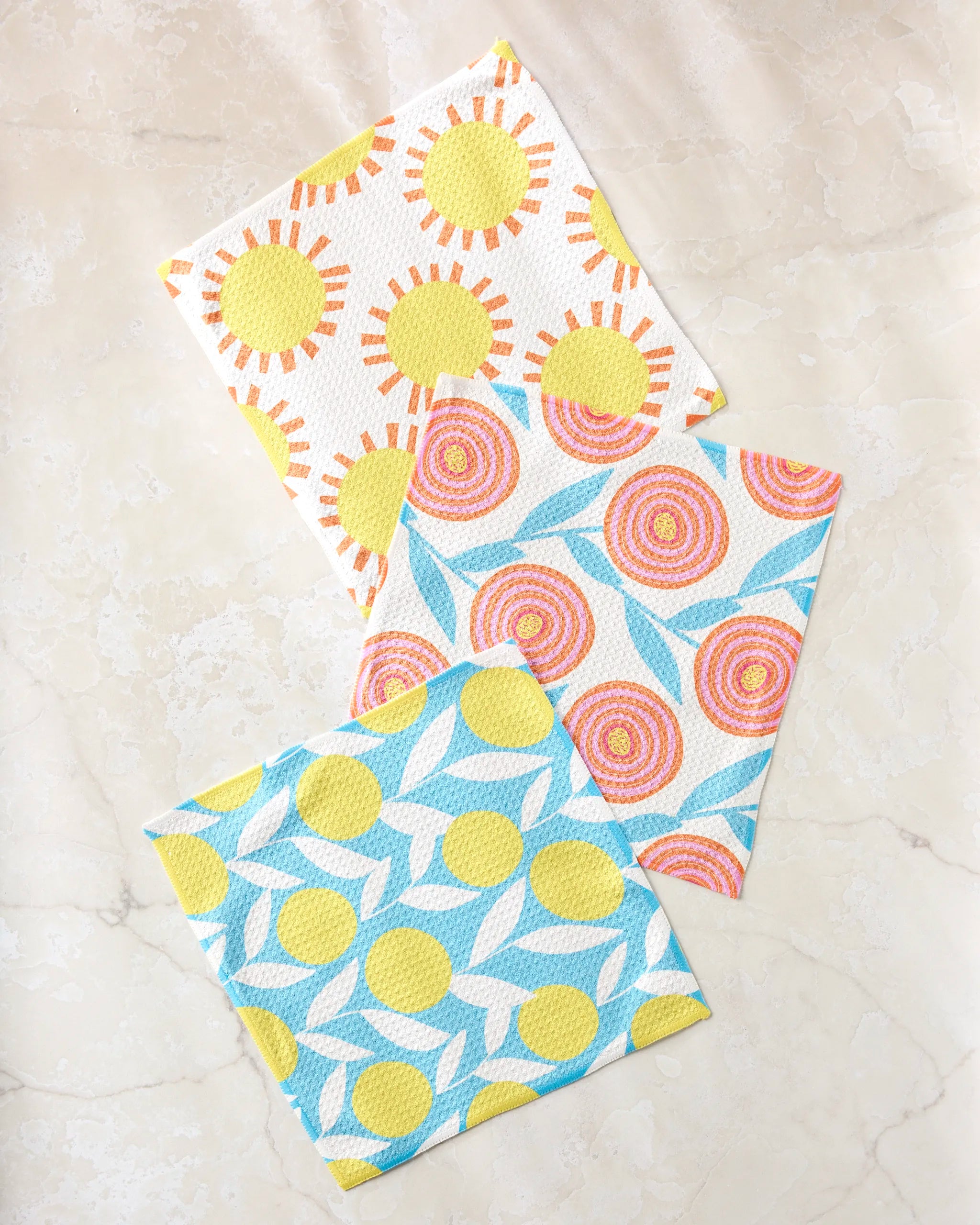 The Lizzie Clark Parade dishcloth set features three super-absorbent, recycled-material towels with sun and floral patterns, combining vibrant style and sustainability on a light marble surface.