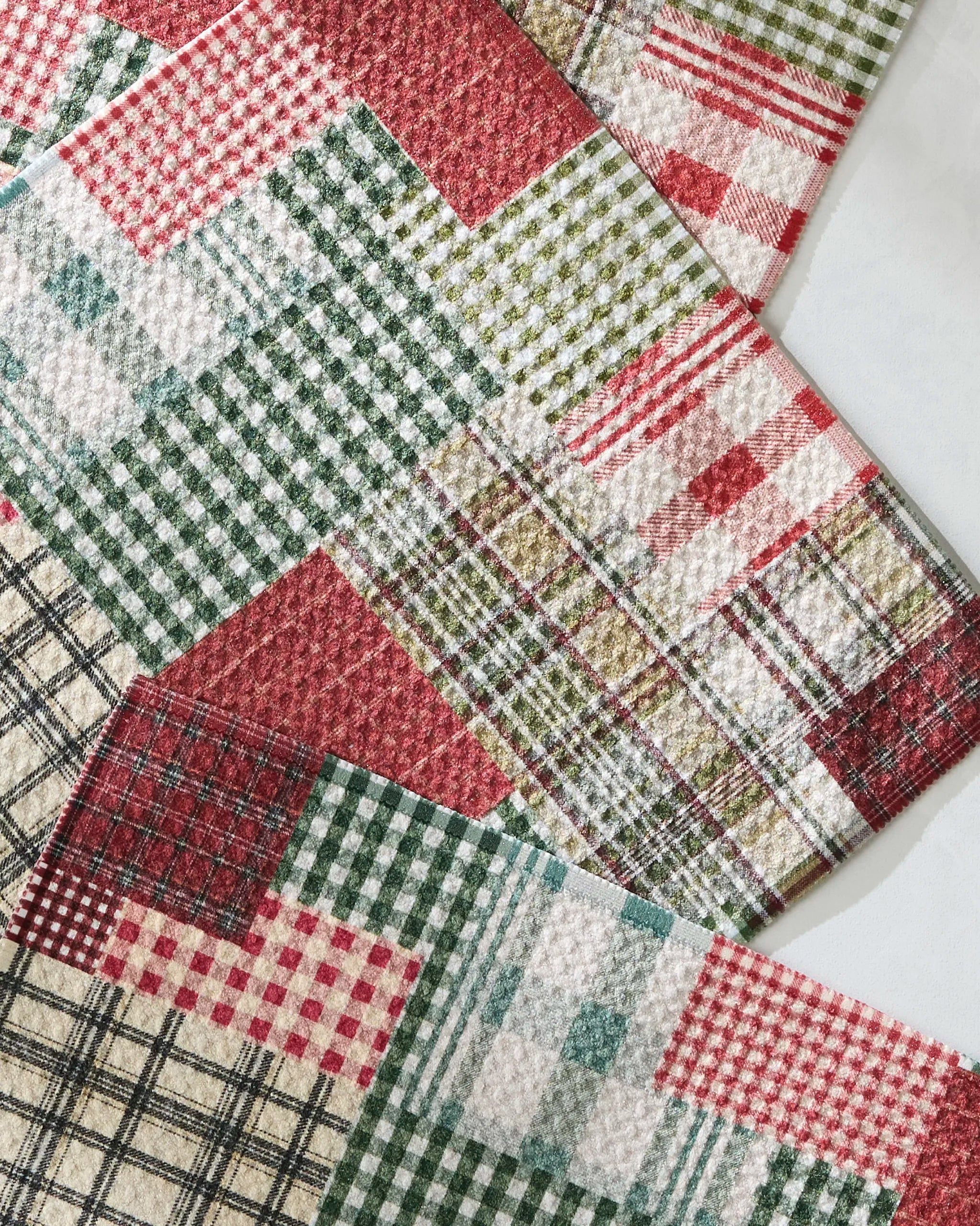 Patchly dishcloths by Geometry feature absorbent, textured fabric surfaces in red, green, and cream plaid, check, and gingham patterns, arranged in a patchwork style.