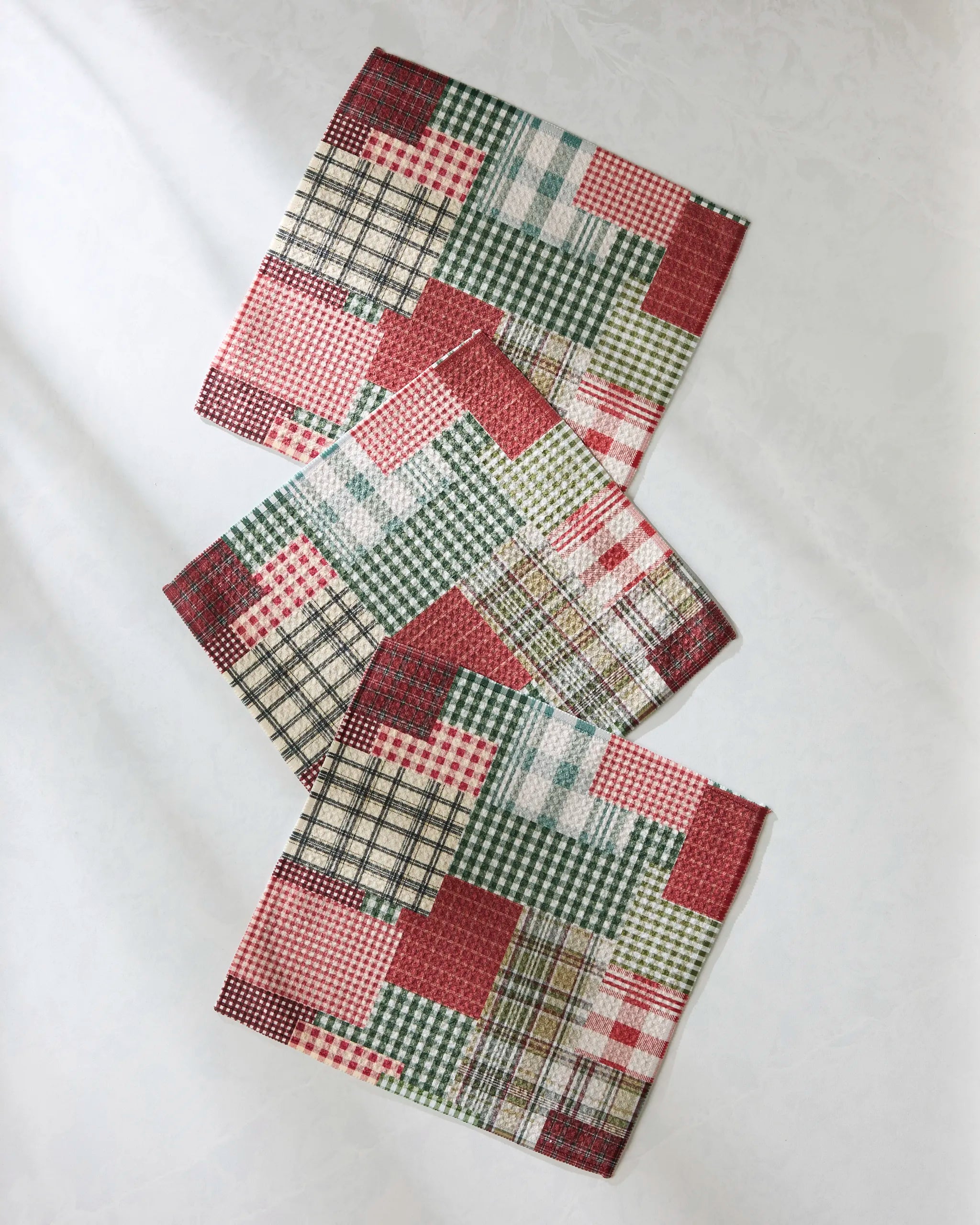 Three Patchly napkins by Geometry, featuring a patchwork of red, green, cream, and black plaid and checkered patterns, are arranged diagonally on a light surface—perfect as charming, absorbent dishcloths for your kitchen.
