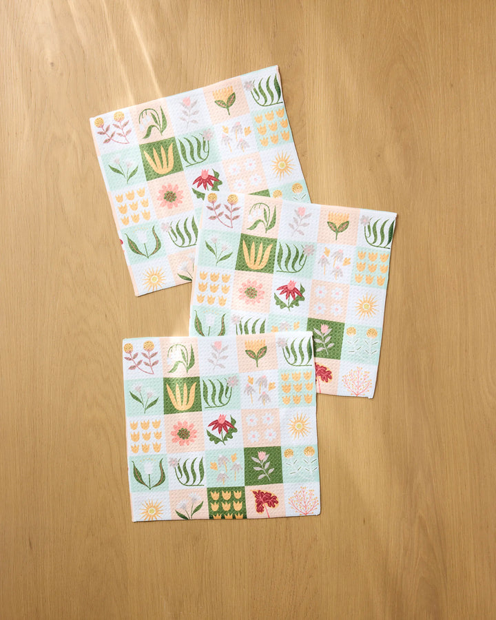 Four Patchwork Garden kitchen towels by Rachel Parker, featuring colorful grid-like patterns of flowers and leaves in pastel and green shades, overlap on a light wooden surface.
