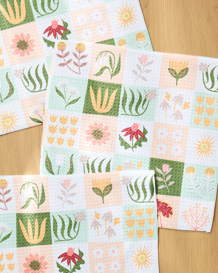 Four Patchwork Garden dishcloths by Rachel Parker, featuring pastel checkered florals in pink, green, yellow, and peach with flowers and sun motifs, are displayed on a light wooden surface.