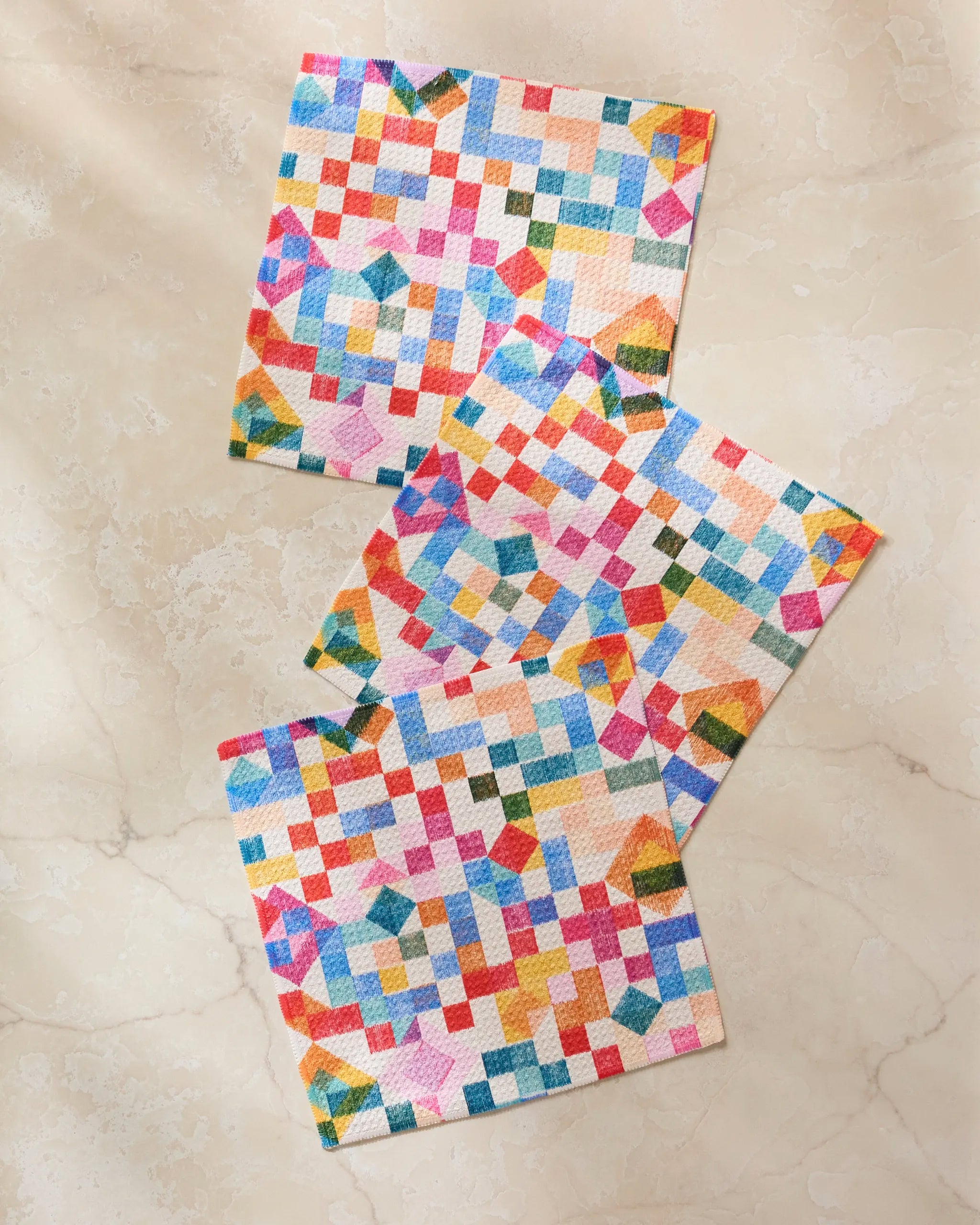 Three colorful Patchwork Geo dishtowels by Rachel Parker, featuring super-absorbent geometric quilt designs in red, blue, yellow, and green, are arranged diagonally on a light beige marble surface.
