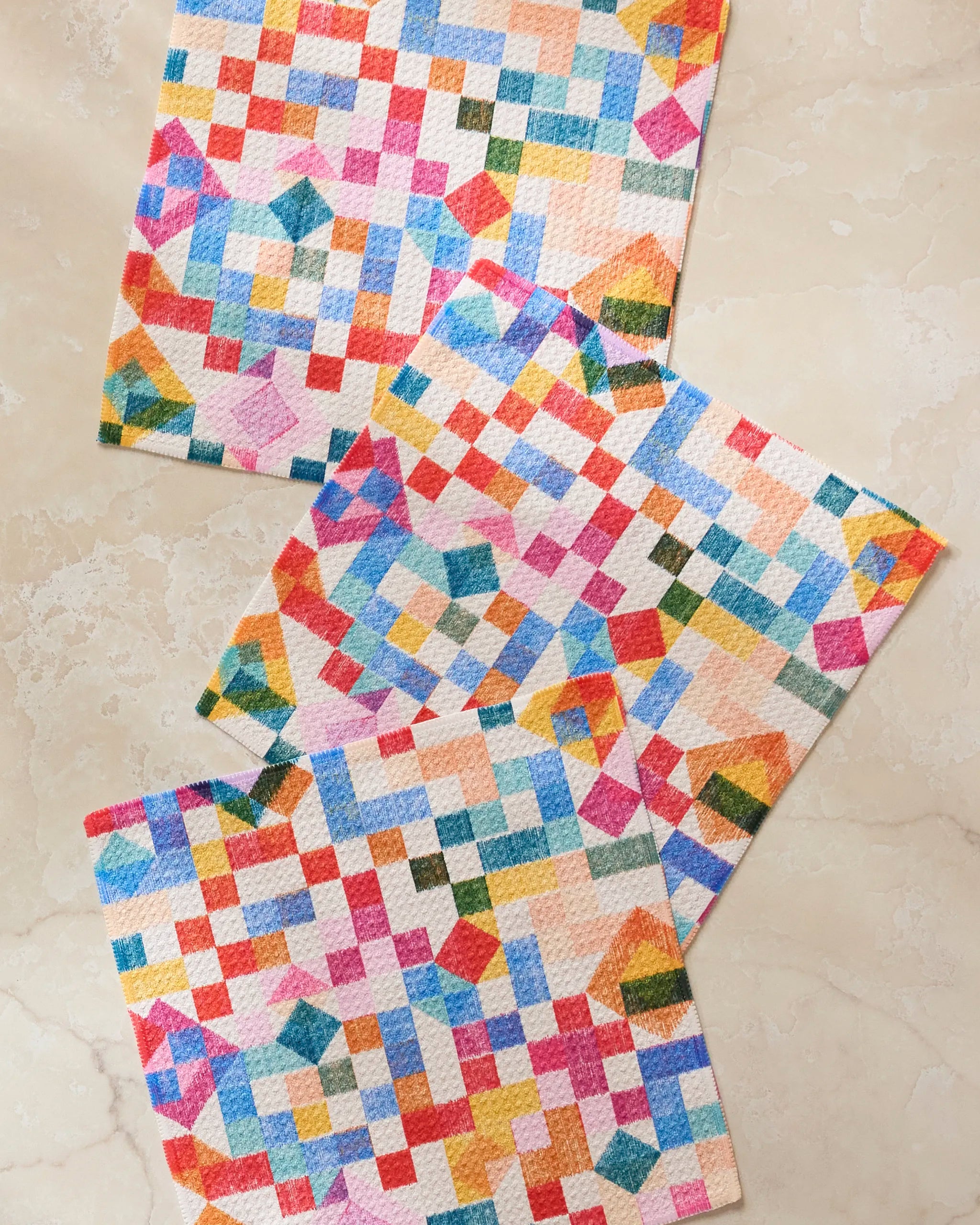 The Patchwork Geo dishcloth set by Rachel Parker features three colorful cloths with geometric patterns in red, blue, yellow, green, and pink, arranged on a beige marble surface—crafted from recycled materials.