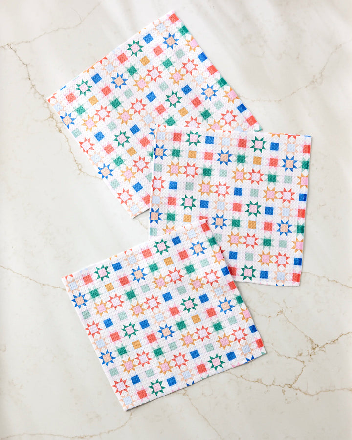 The Patchwork Picnic dishcloth set by Brittany Jepsen features three square cloths with vibrant geometric star and square patterns in orange, green, blue, and red—made from recycled materials and displayed on a marble-like surface.
