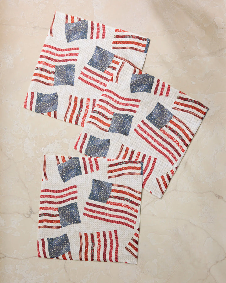 Geometry’s Patchwork Pride napkins, featuring a bold American flag motif, are arranged on a beige marble surface—perfect to pair with your favorite recycled dishcloths for easy and stylish kitchen cleanup.