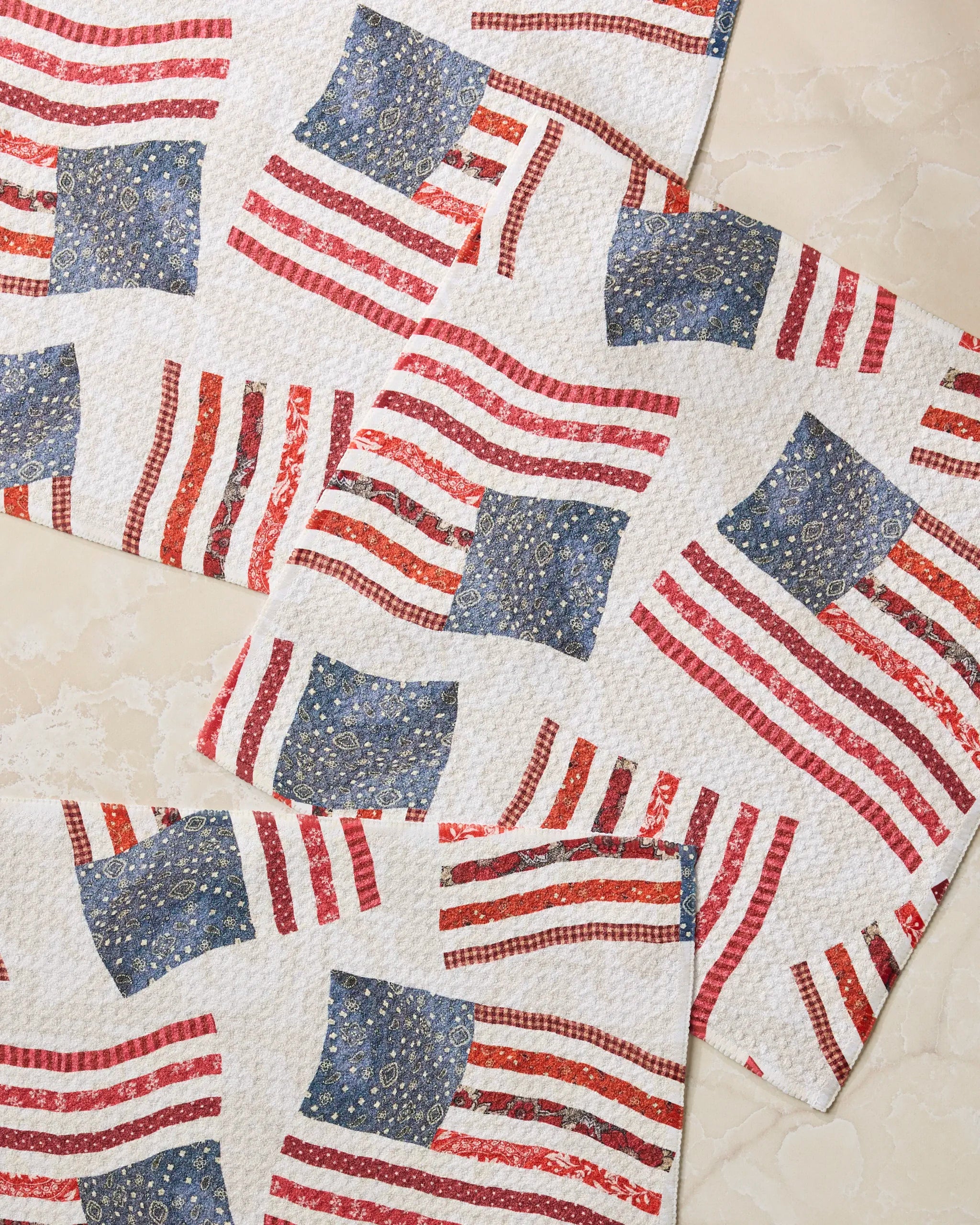 Three Patchwork Pride placemats by Geometry, featuring abstract American flag designs with blue stars and red stripes, look great on a marble surface and pair perfectly with our dishcloth set for stylish, convenient kitchen setups.