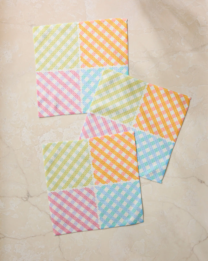 Three Patio Check square napkins by Geometry, featuring a colorful checkered pattern in pastel yellow, orange, blue, pink, and green, are displayed on marble—a vibrant set that adds brightness to any kitchen.
