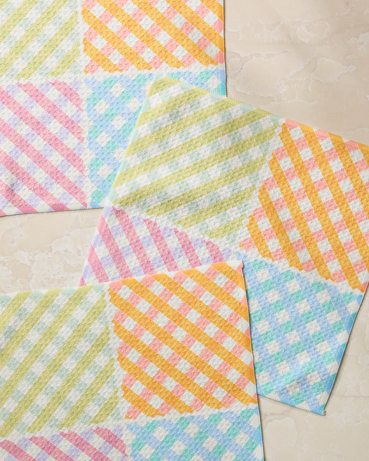 Three pastel cloths from Geometry’s Patio Check Dishcloth Set, with diagonal checkered pink, blue, green, and orange squares, are displayed on a light beige marble surface.