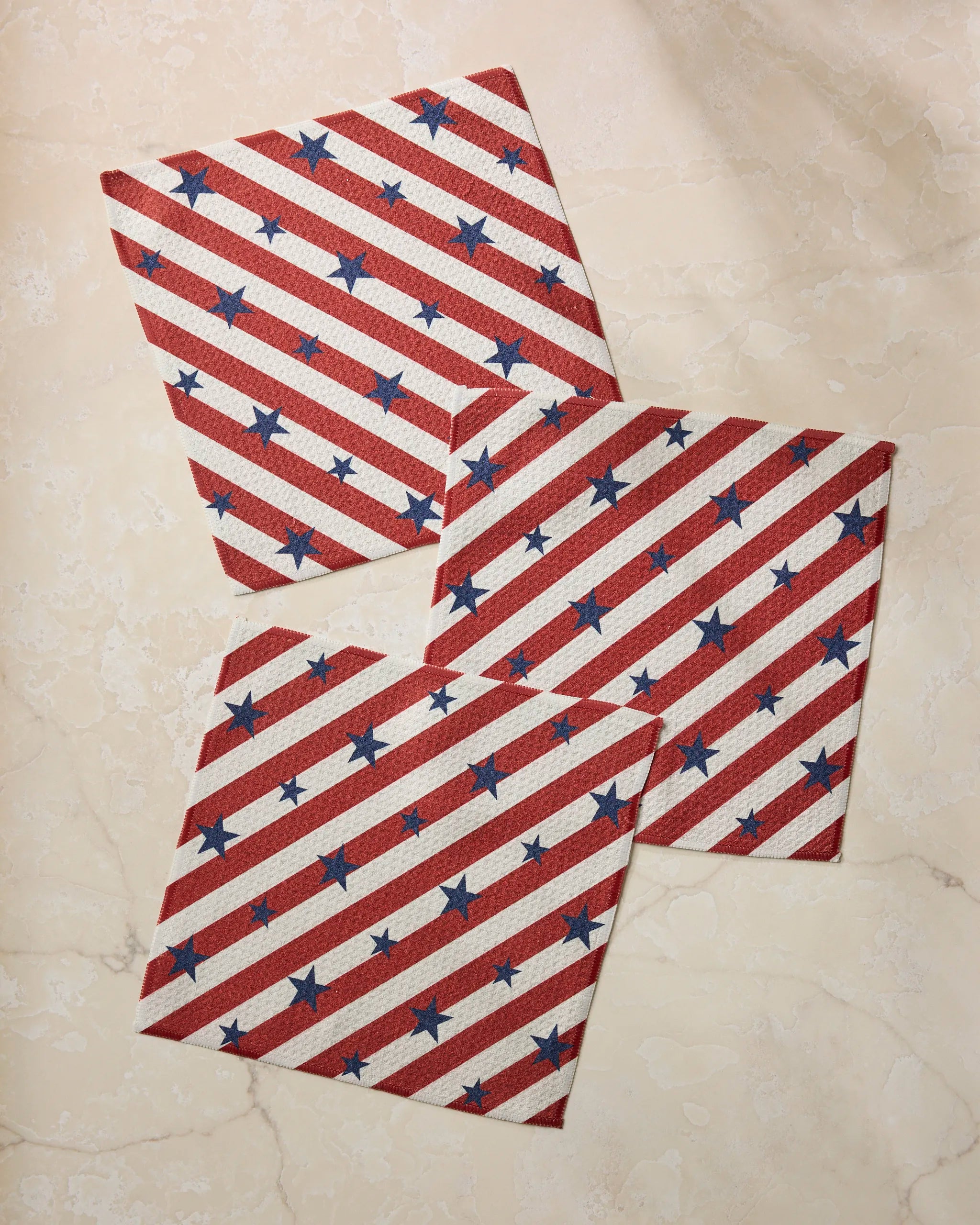 The Geometry Patriotic Stripe set features three square napkins made from recycled materials, with red and white diagonal stripes and blue stars, arranged on a light marble surface.