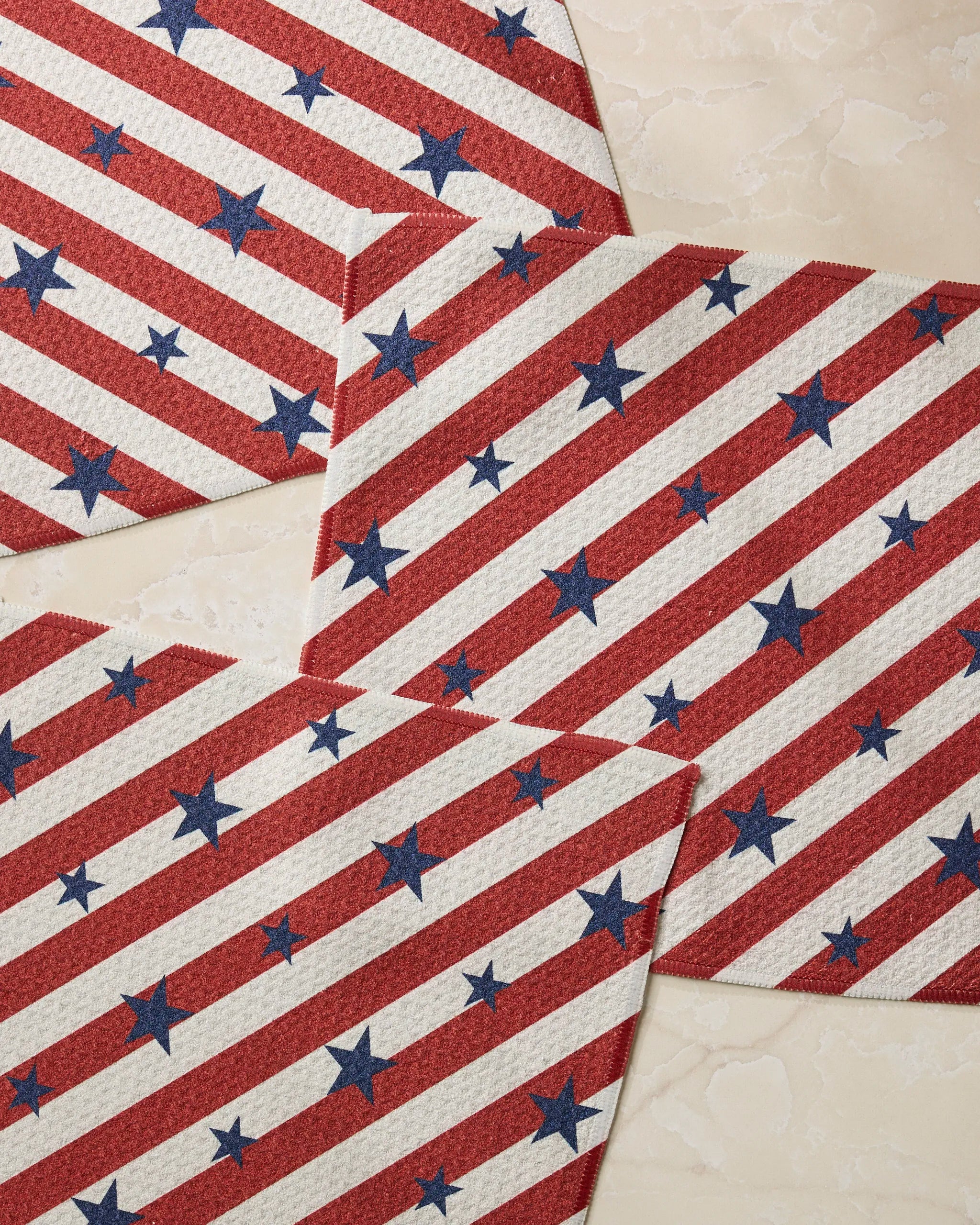 Patriotic Stripe placemats by Geometry, featuring red and white stripes with blue stars in an American flag-inspired design, are arranged overlapping on a light beige textured surface and made from recycled materials.