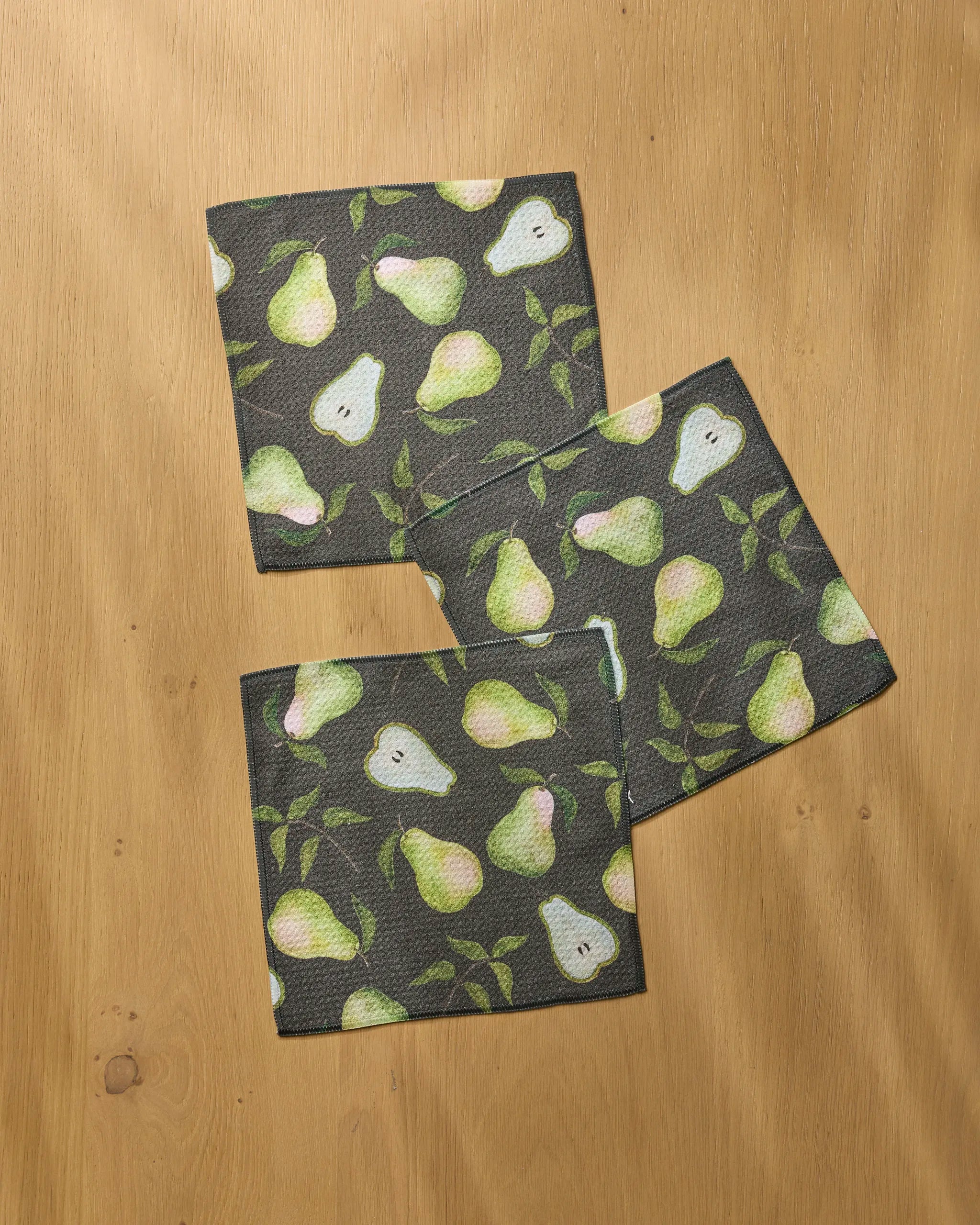 Three dark-colored Geometry Pearwood napkins are arranged on a wooden surface. Made from recycled materials, each napkin features green and yellow pears with scattered green leaves.
