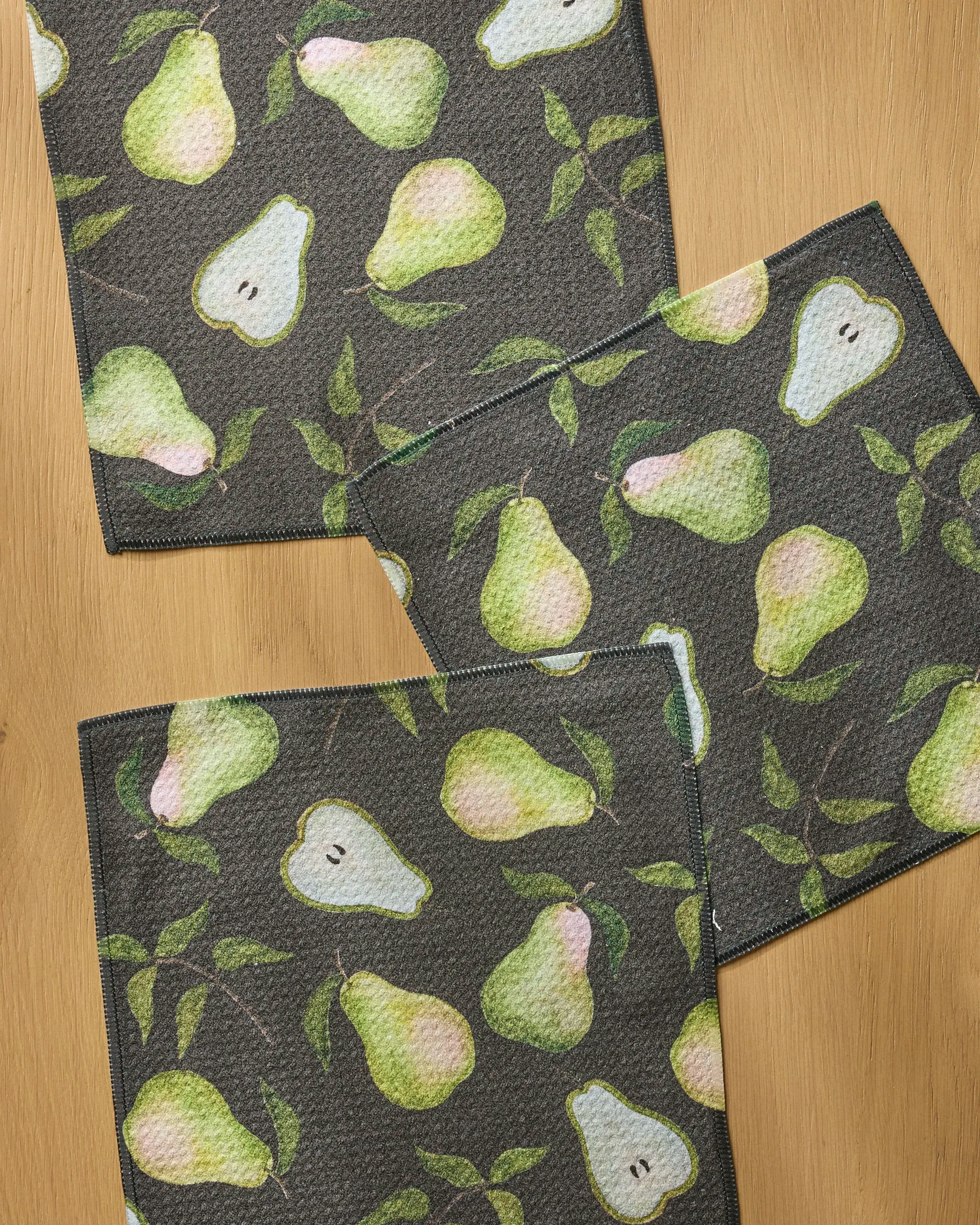 The Geometry Pearwood set includes three super-absorbent dishcloths with a watercolor pear design—whole and sliced pears with green leaves. Two cloths have dark backgrounds and one is light, all arranged on a wooden surface.
