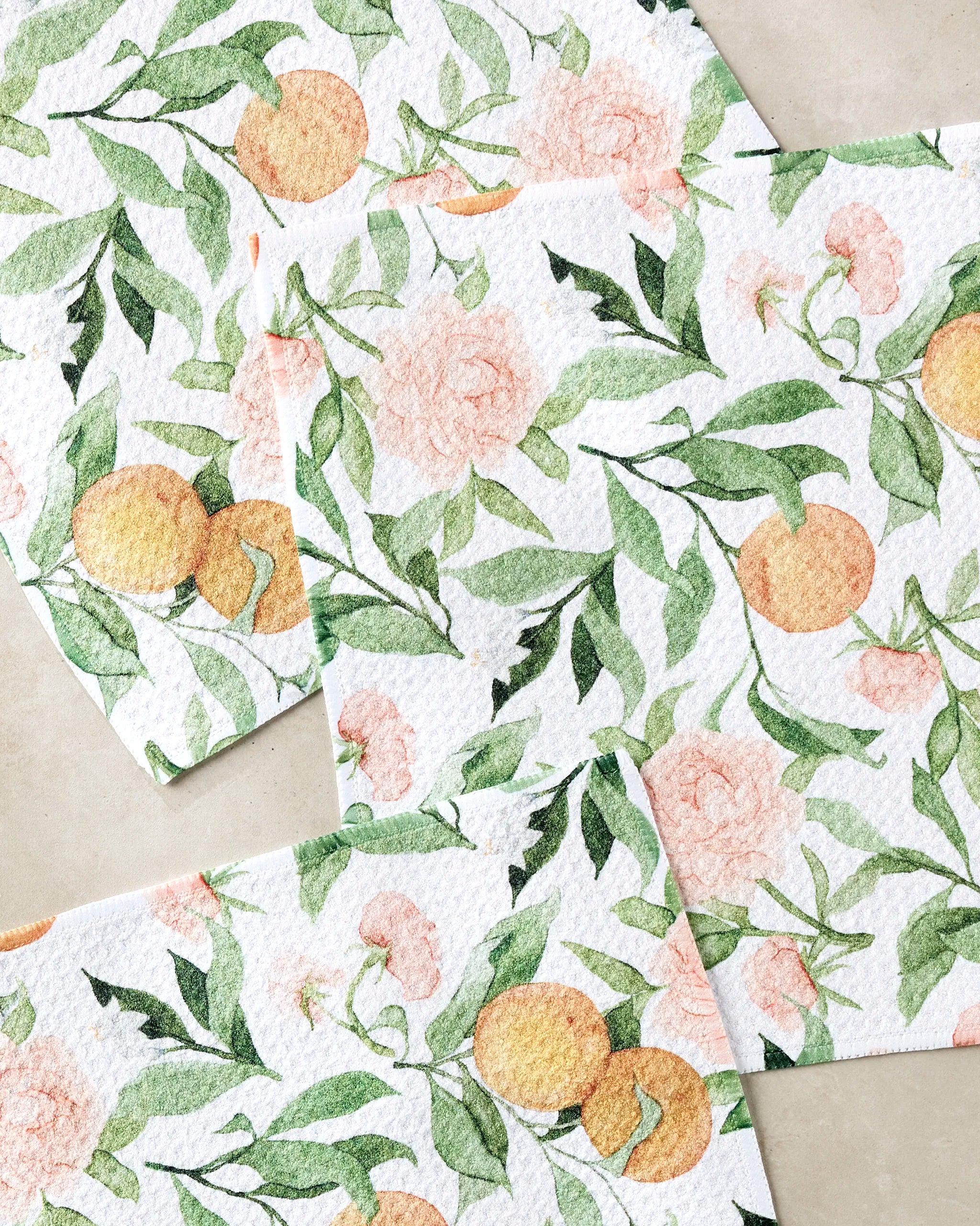 Three overlapping "Peony For Your Thoughts" paper napkins by Aubrey Fairchild, made from recycled materials and adorned with watercolor oranges, green leaves, and soft pink flowers, are arranged on a light surface.