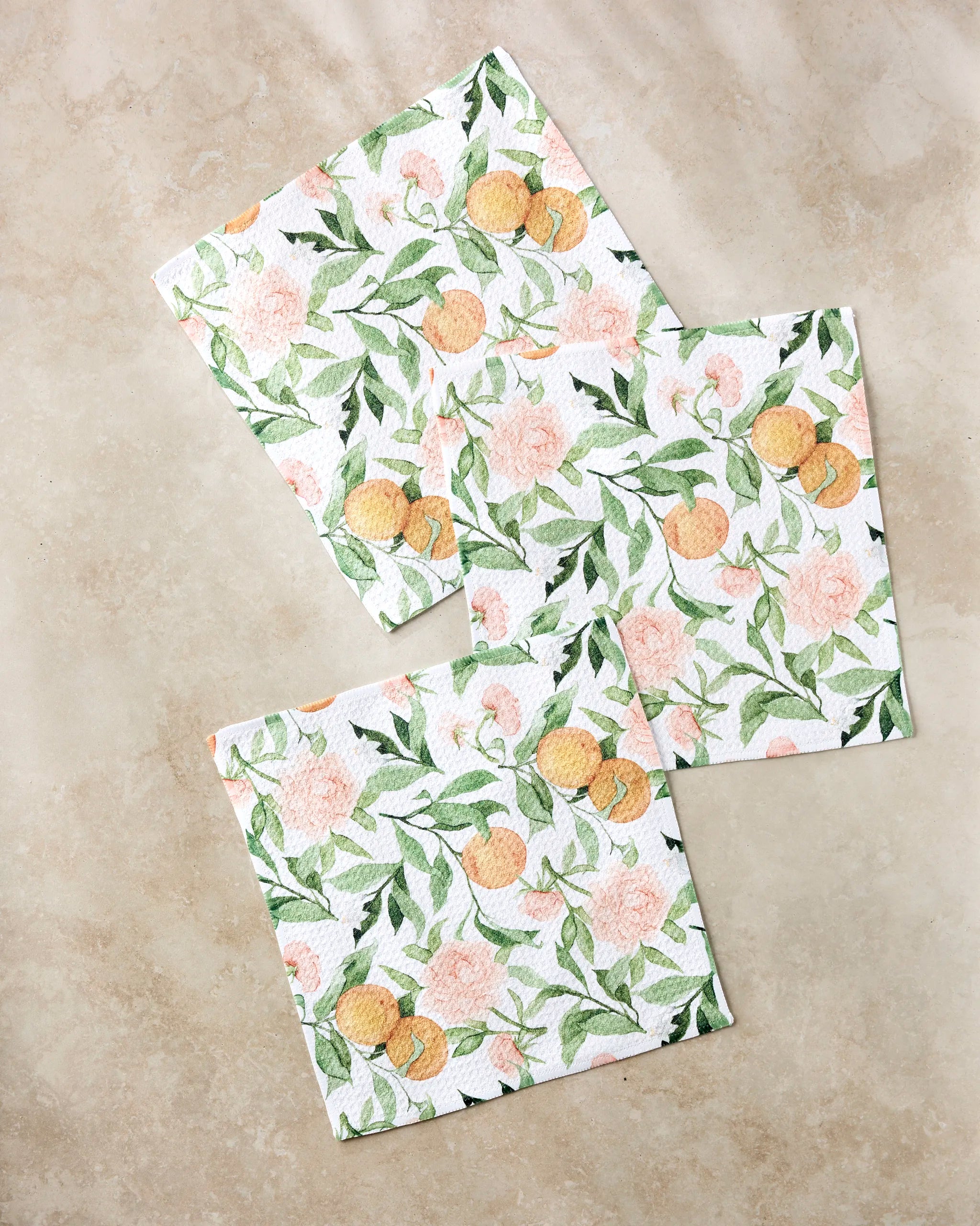 Three “Peony For Your Thoughts” square napkins by Aubrey Fairchild, crafted from recycled materials, display green leaves, peach flowers, and small orange fruits. They’re beautifully arranged on a light beige marble surface.
