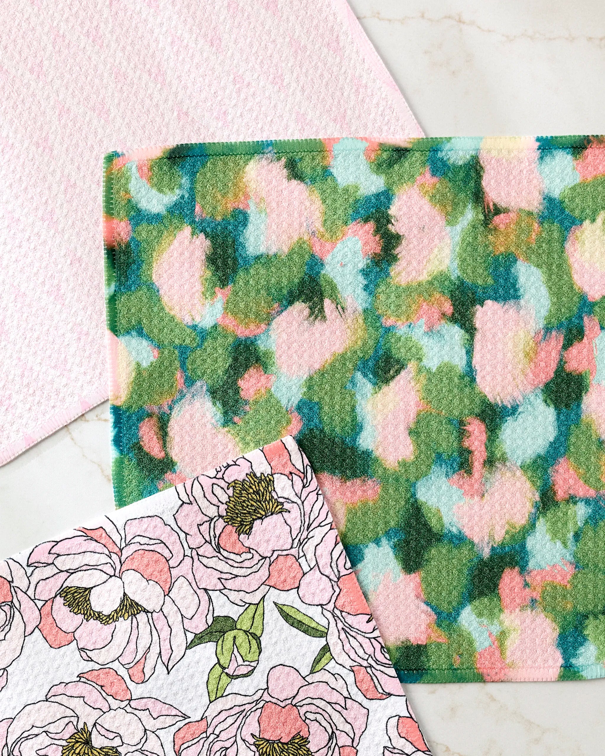 The Shelley Smith Peony Paradise dishcloth set includes three super-absorbent towels: a pink textured towel, a colorful abstract print, and a white towel with hand-drawn pink peonies—ideal for brightening any kitchen.